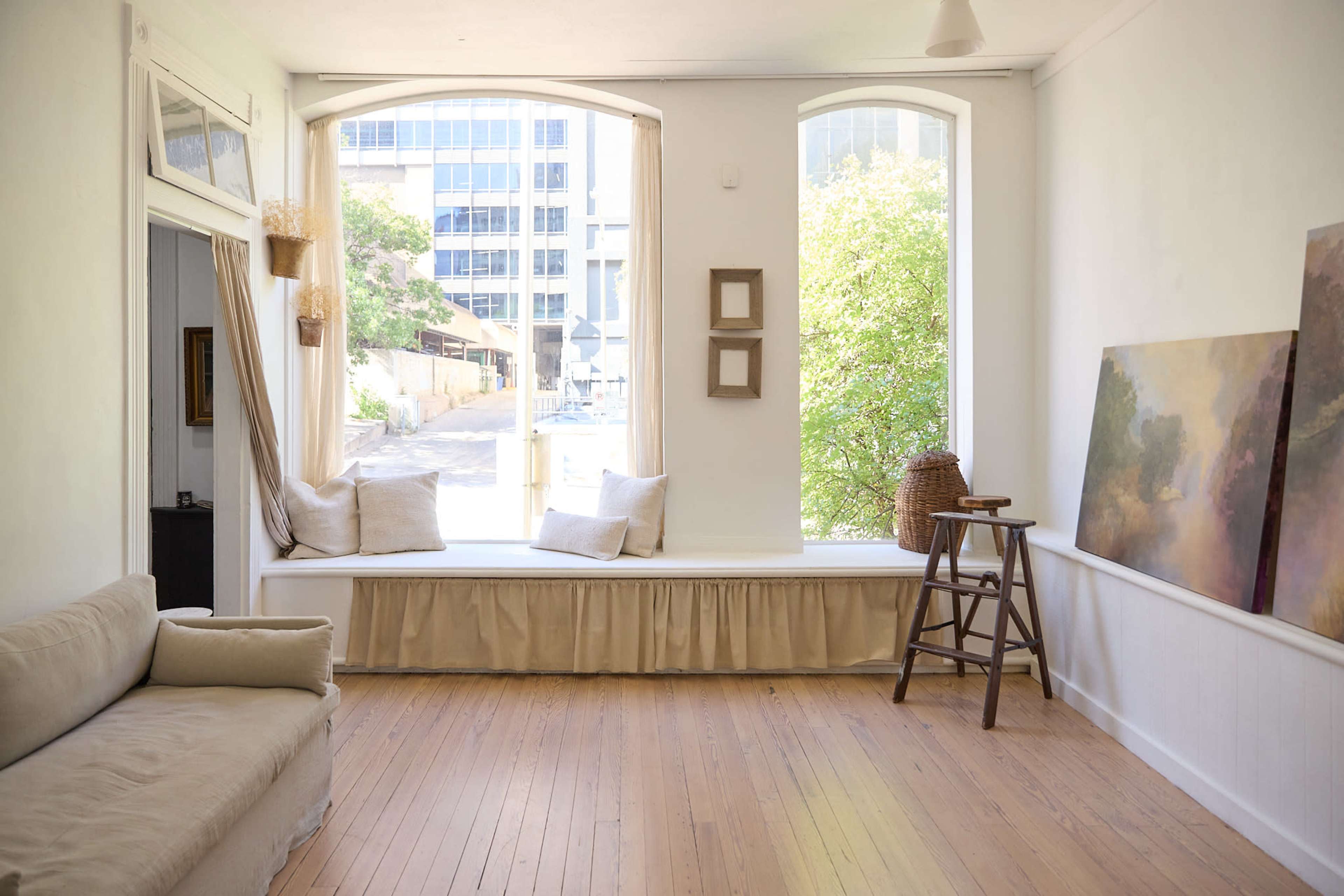 The image shows a bright, minimalist interior space with a window seat, two paintings, and light wooden flooring.