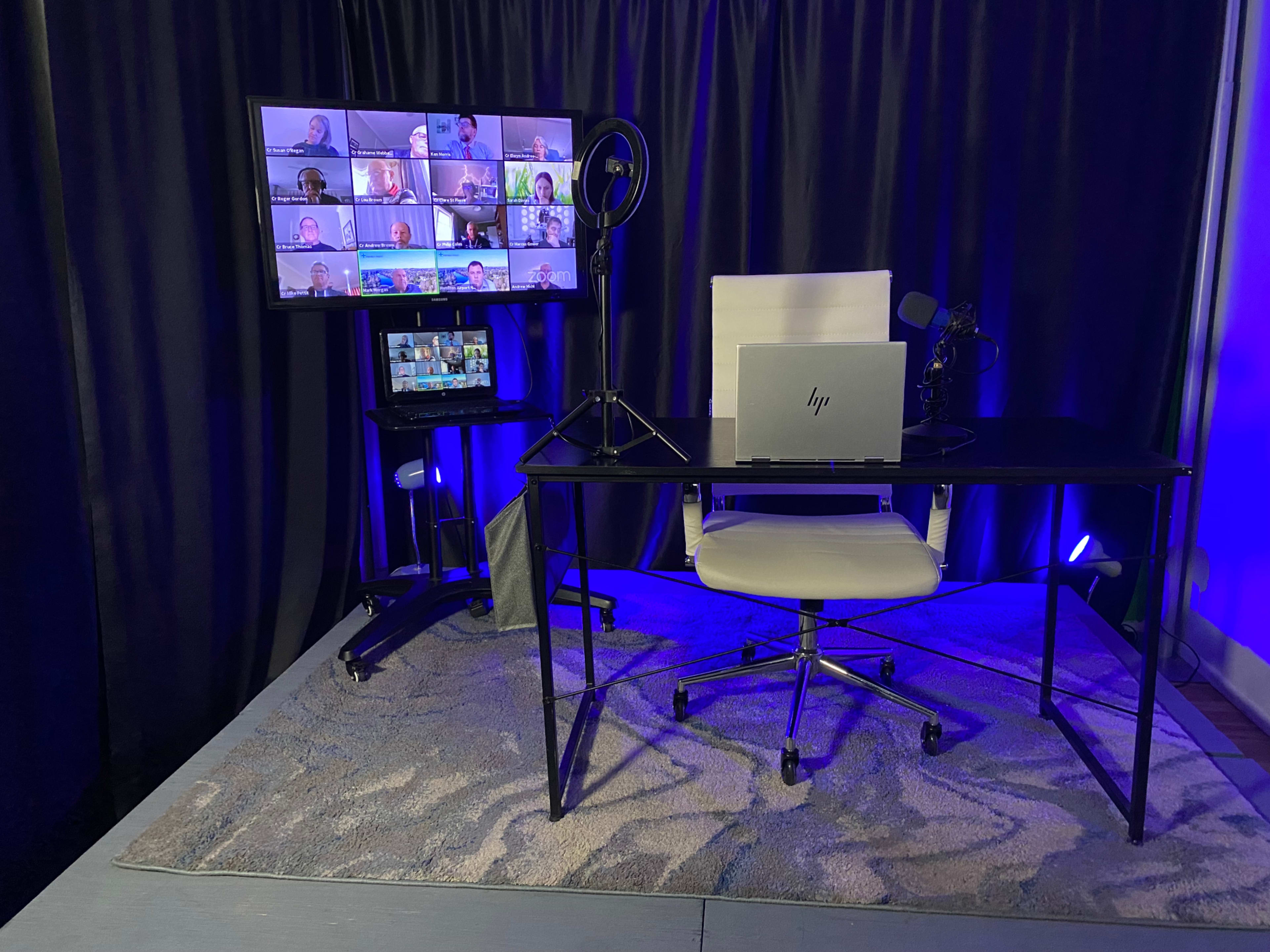 A modern workspace set up for video conferencing, featuring a desk with a laptop, microphones, and multiple screens displaying participants.