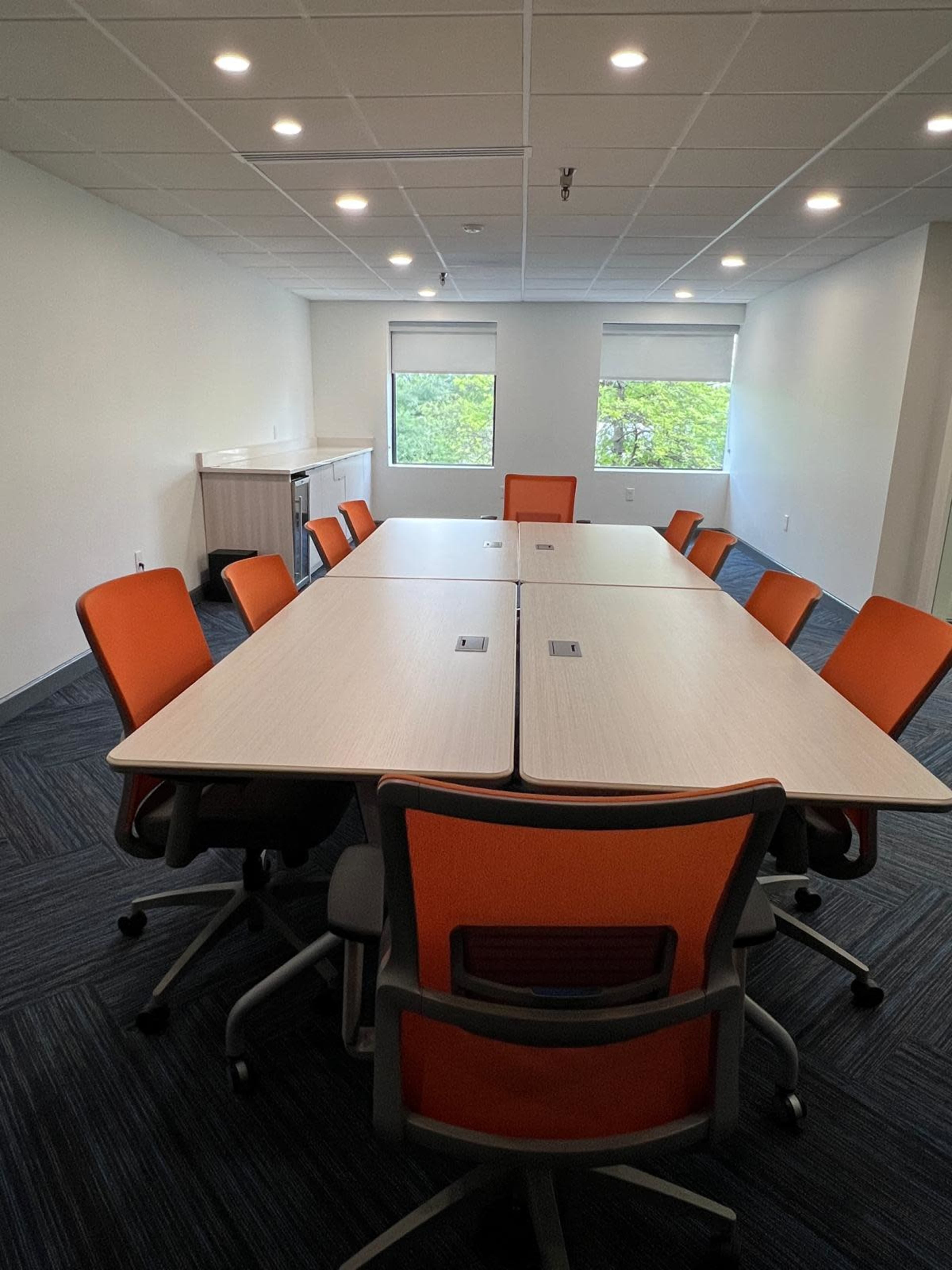 A spacious meeting room features a large table surrounded by orange chairs and windows letting in natural light.