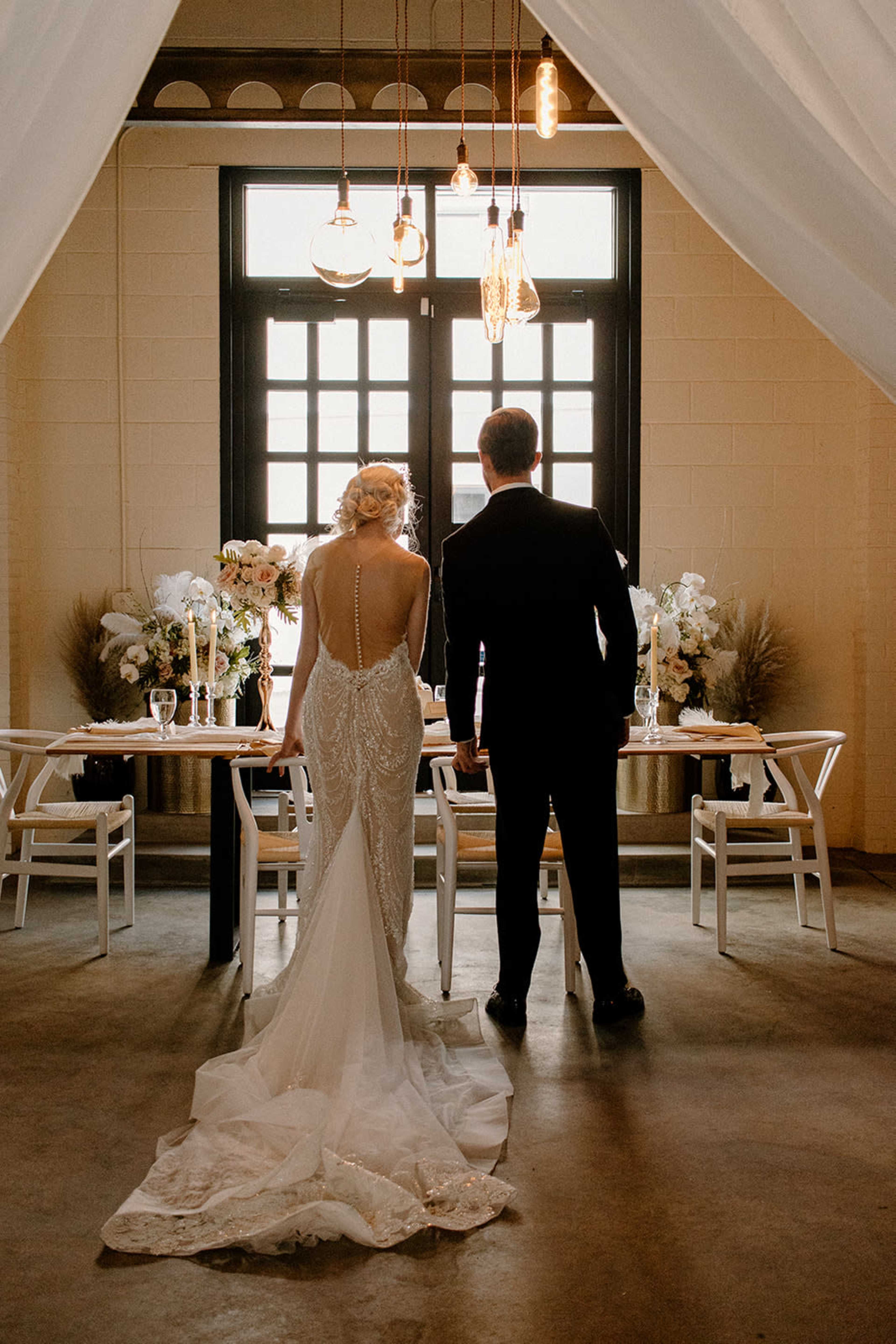 A bride and groom stand together, facing a elegantly set dining table adorned with floral arrangements and candles, under warm hanging lights in a stylish indoor venue.
