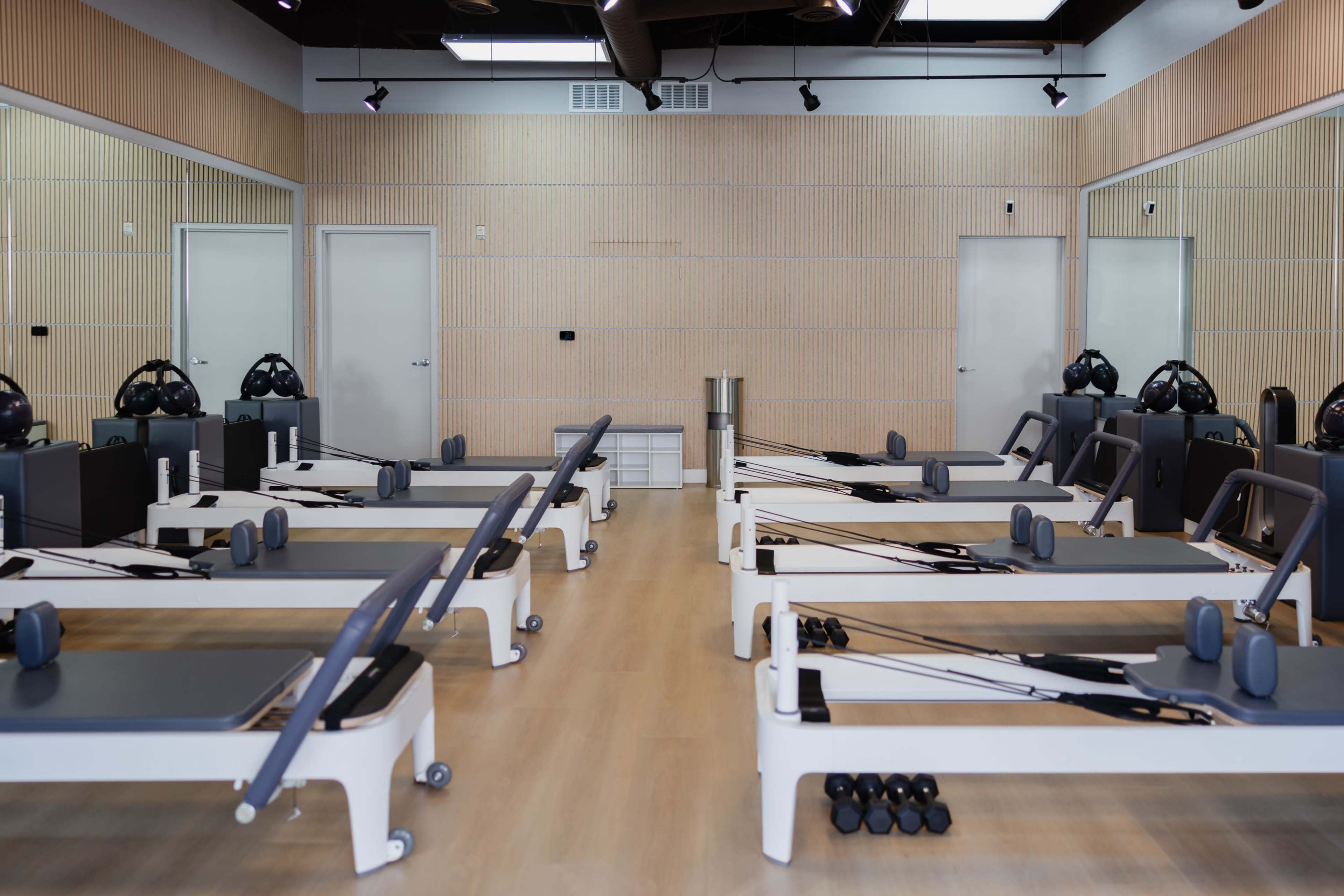 The image shows a spacious pilates studio equipped with several reformer machines and weights, arranged neatly on a light wood floor.