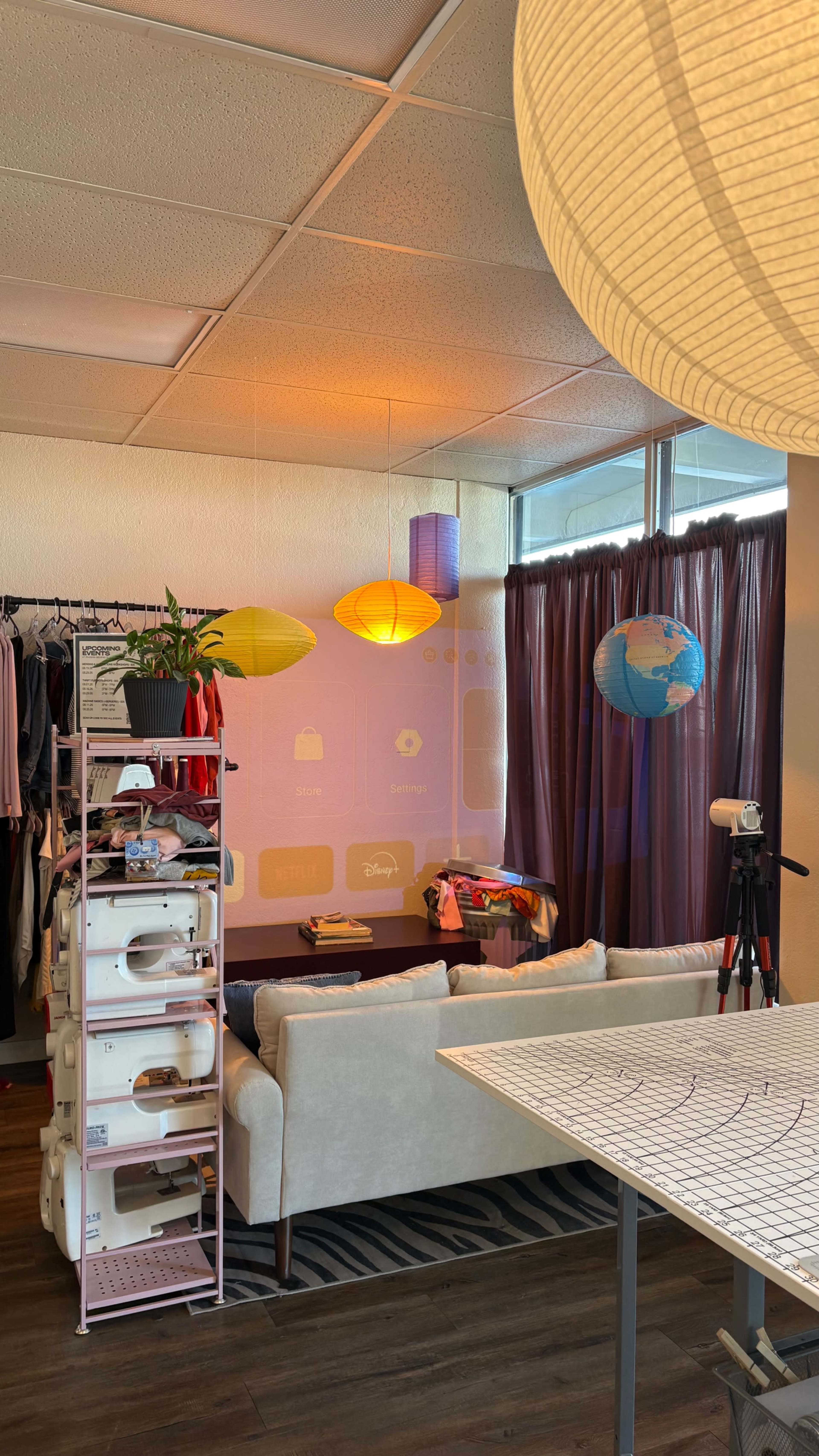 The image shows a cozy room with a light-colored sofa, a rack of clothes, colorful paper lanterns hanging from the ceiling, and a table with a cutting mat.