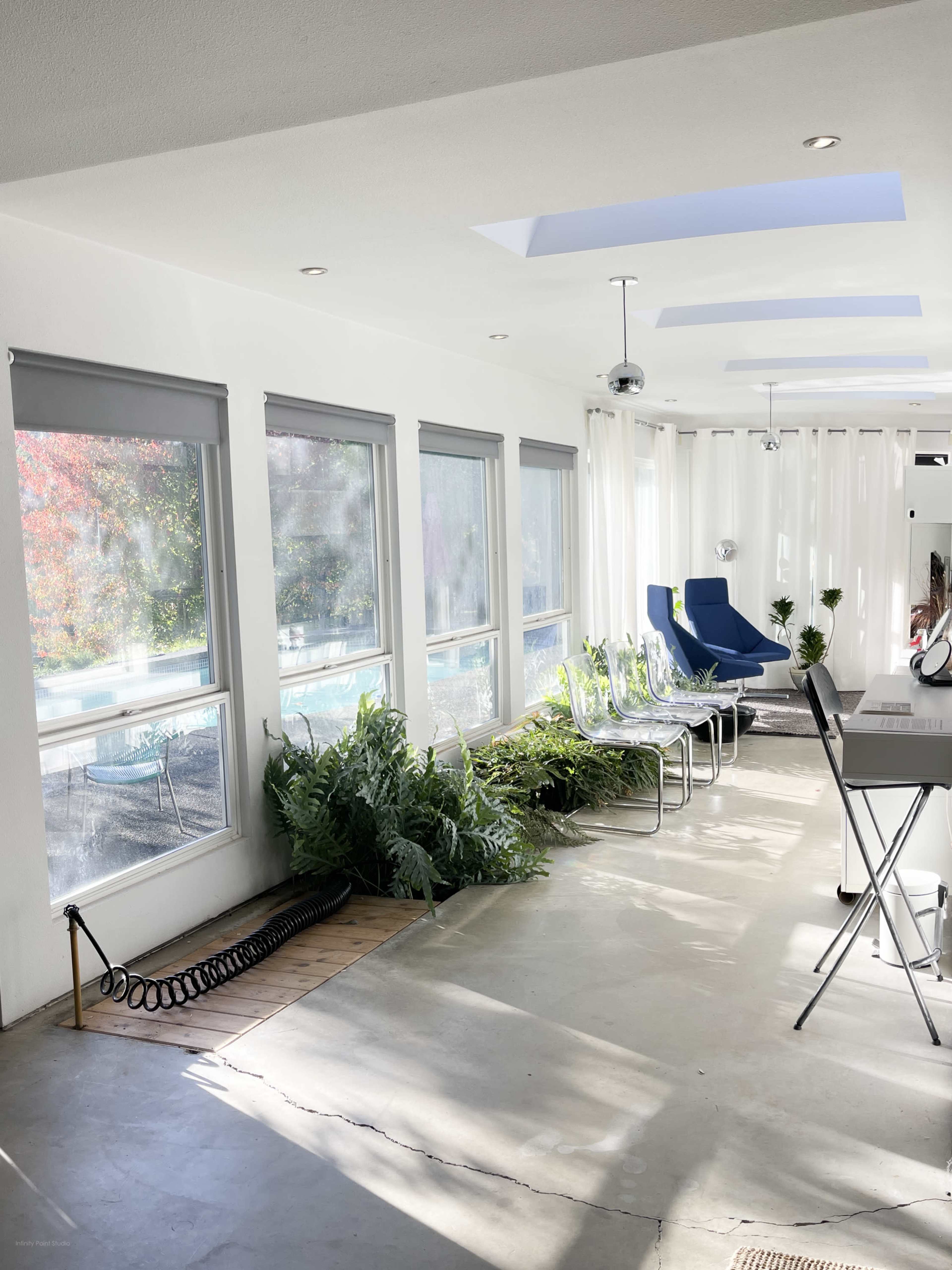 The image shows a spacious, bright room with large windows, sunlight streaming in, metal chairs lined along one side, and greenery placed near the floor.