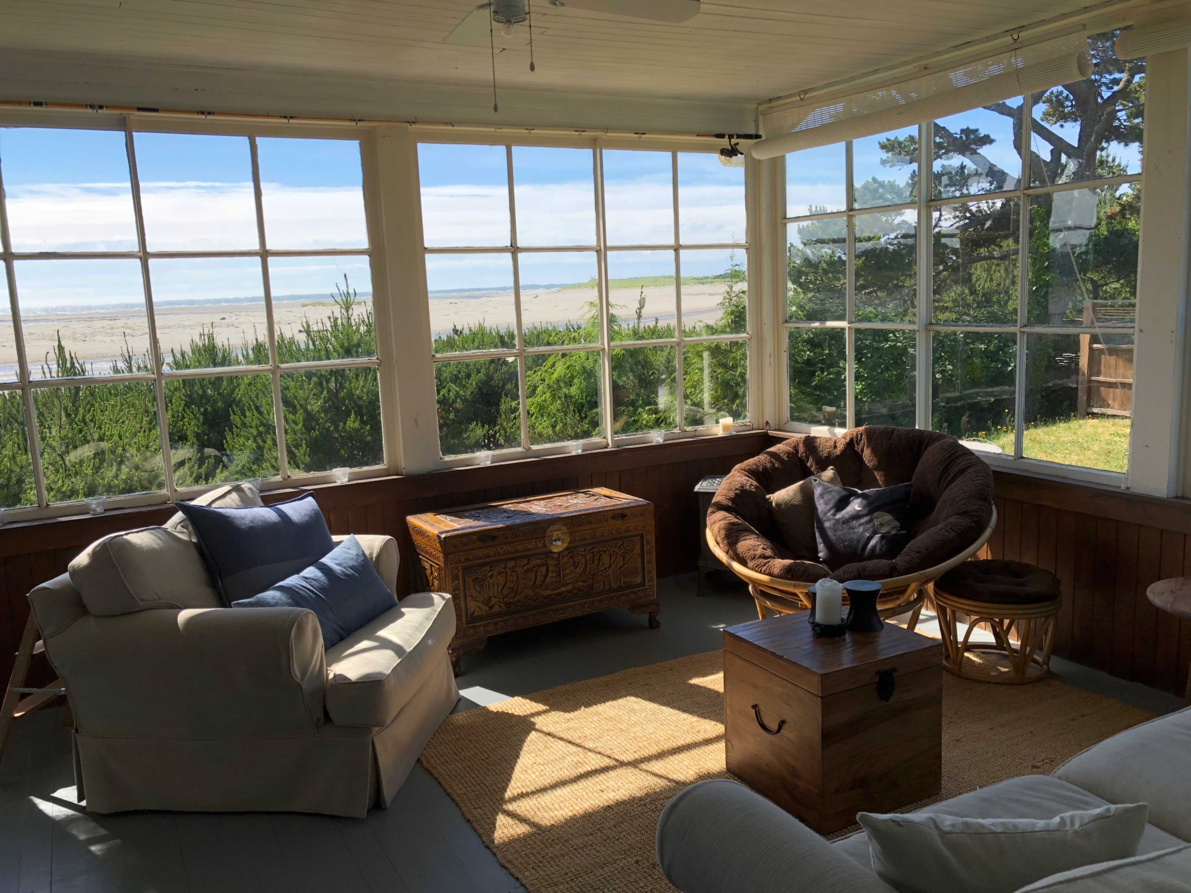 A sunlit, enclosed porch features a pair of sofas, a round chair, and a wooden chest, with large windows displaying a view of the beach and greenery outside.