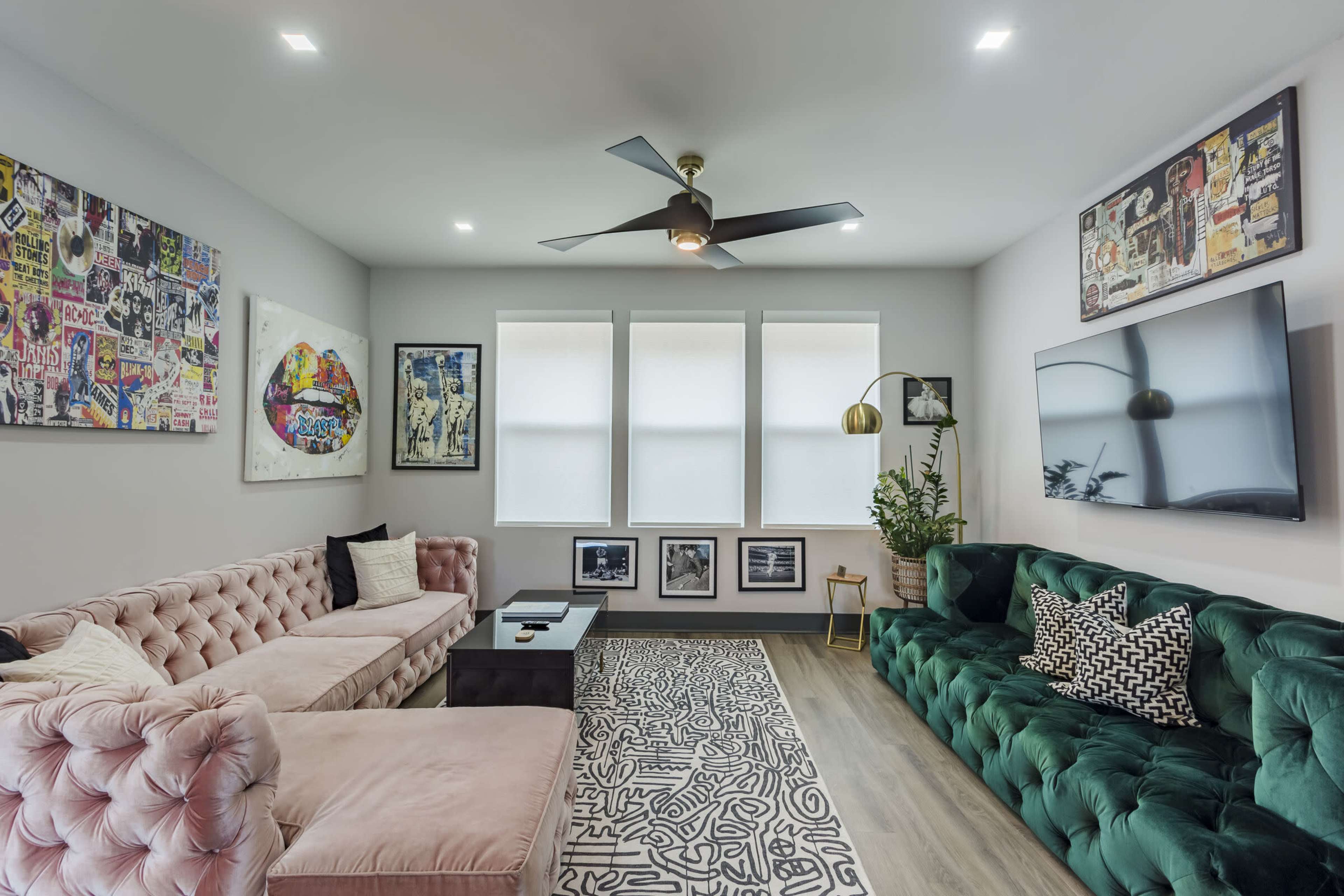 The room features a mix of stylish furniture, including a pink tufted sofa and a green velvet couch, with artwork on the walls and a large television mounted across from them.