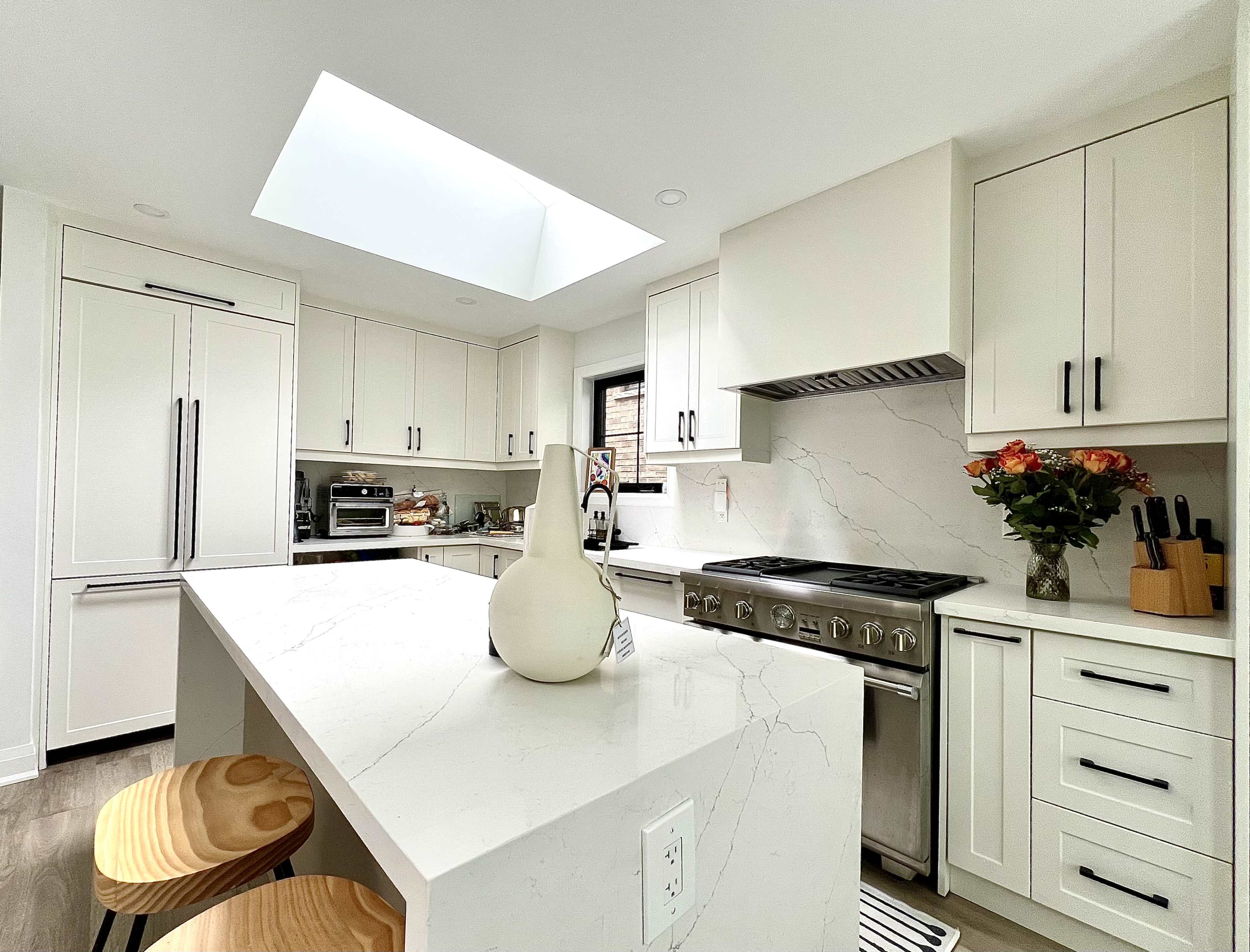 A modern kitchen features white cabinetry, a large island with a marble countertop, stainless steel appliances, and a skylight illuminating the space.
