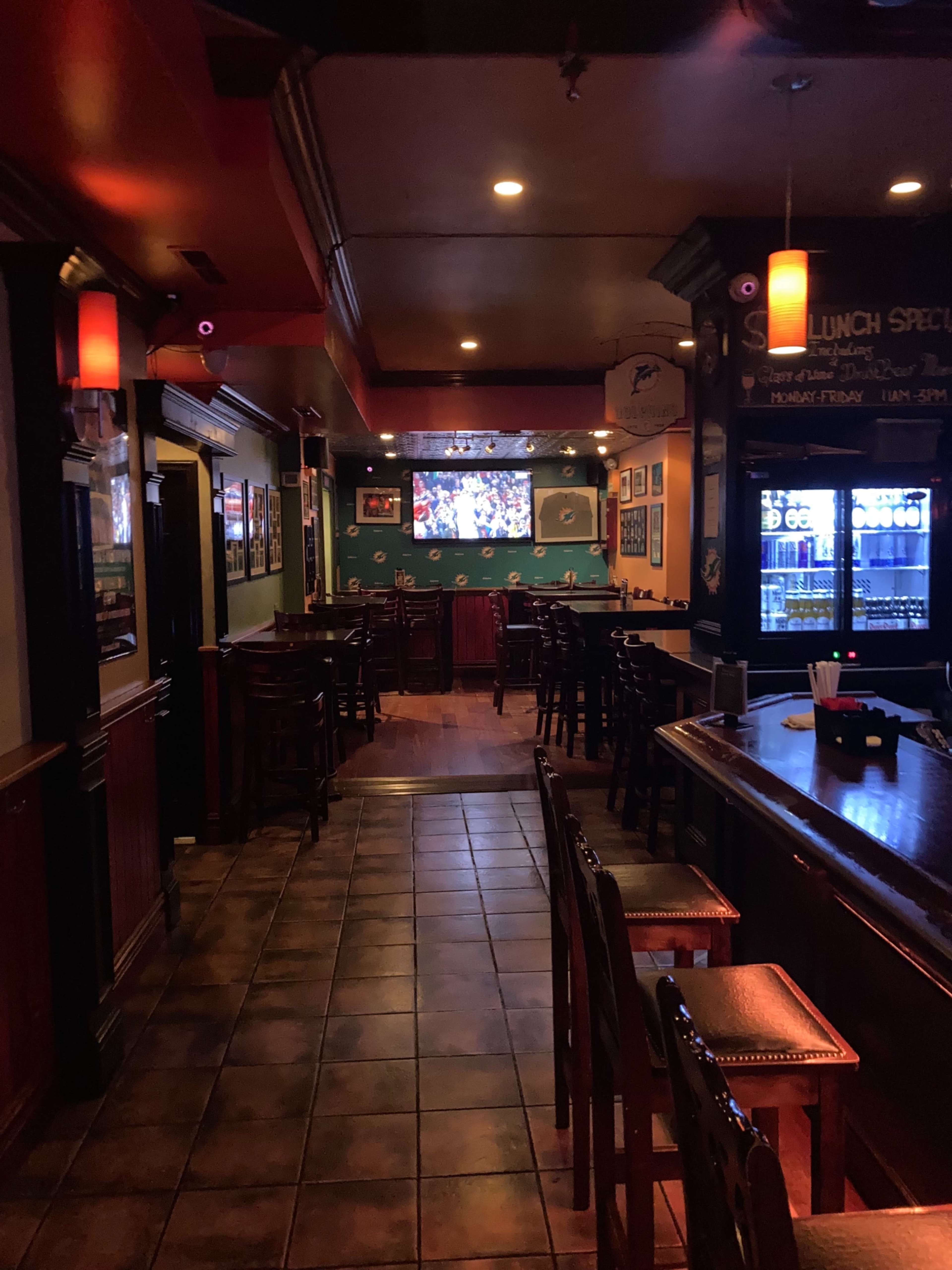 The image shows a dimly lit bar interior with rows of wooden chairs and a television screen displaying a sports event on the wall.