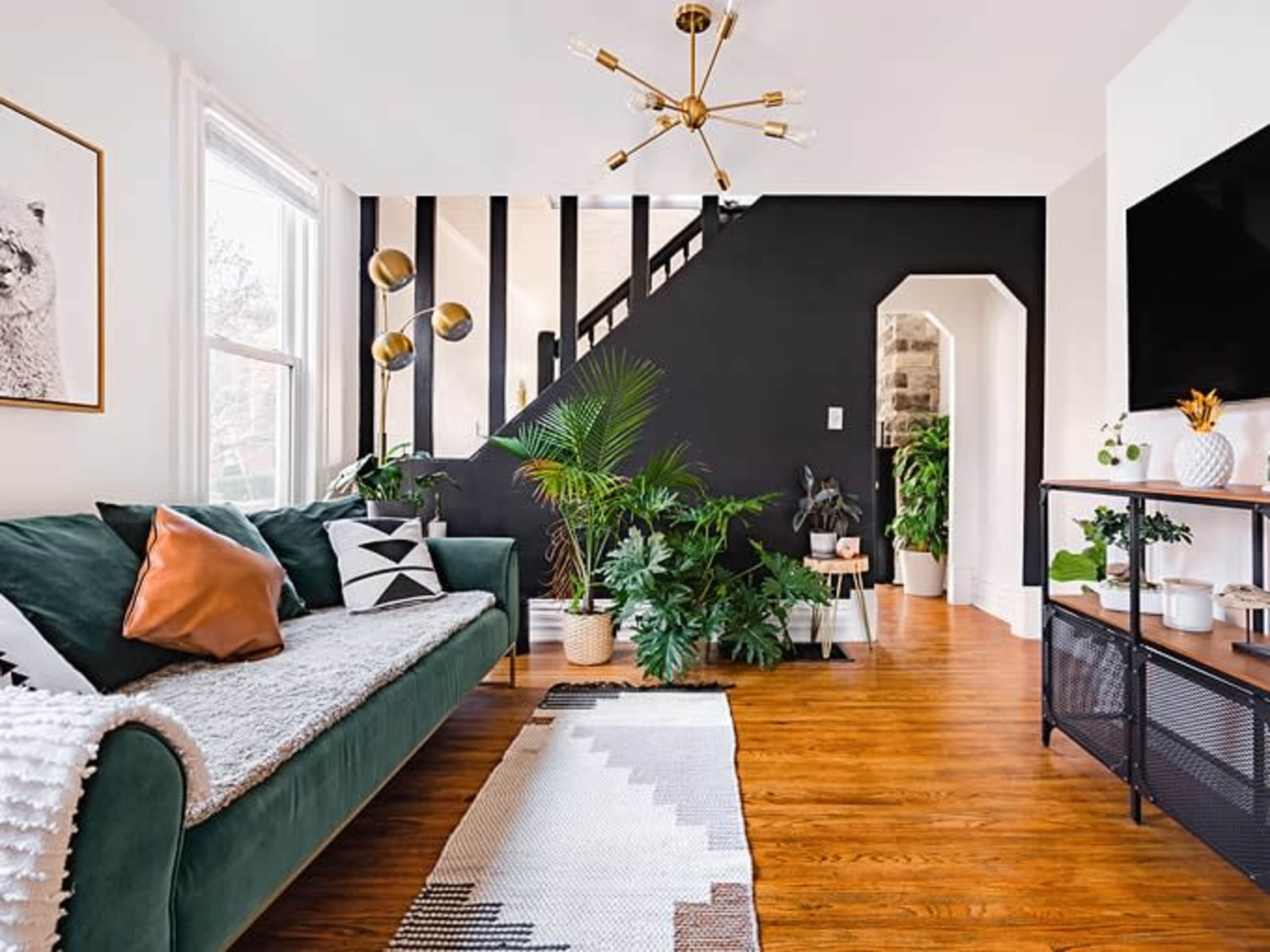 A modern living room featuring a green sofa with decorative pillows, a geometric rug, and a staircase leading to an upper level against a black accent wall, accompanied by various indoor plants and a television setup.