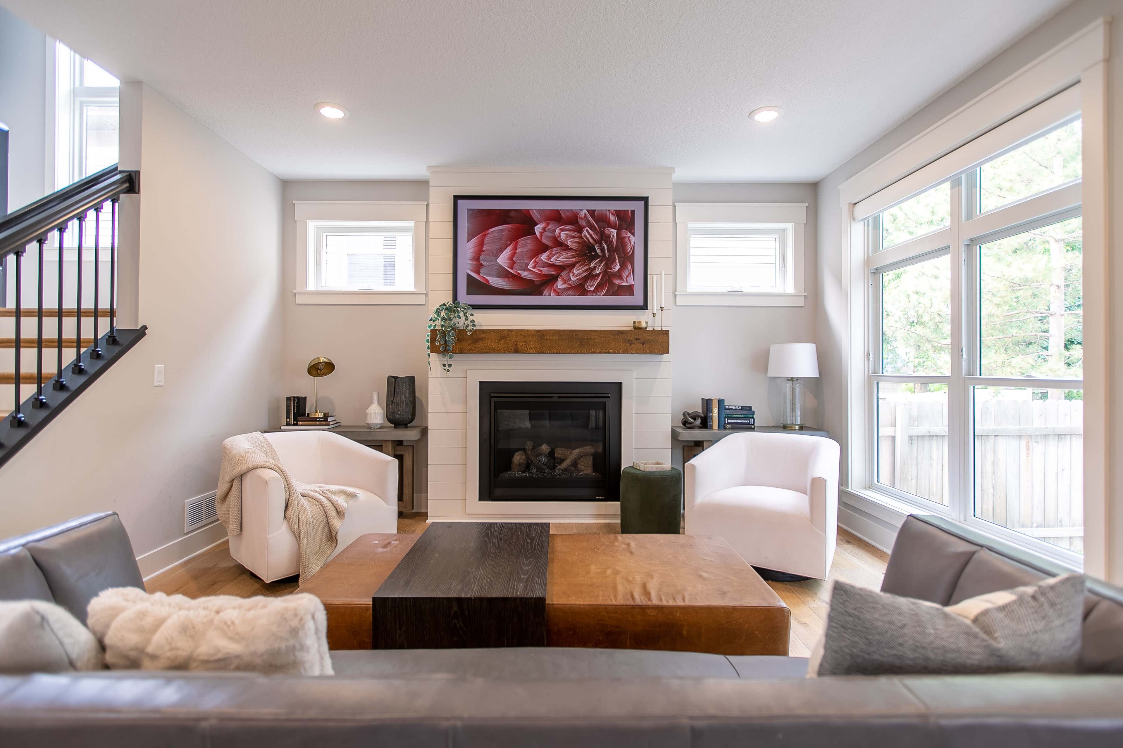 A modern living room featuring a fireplace with a mounted television above it, two armchairs, a wooden coffee table, and large windows allowing natural light.
