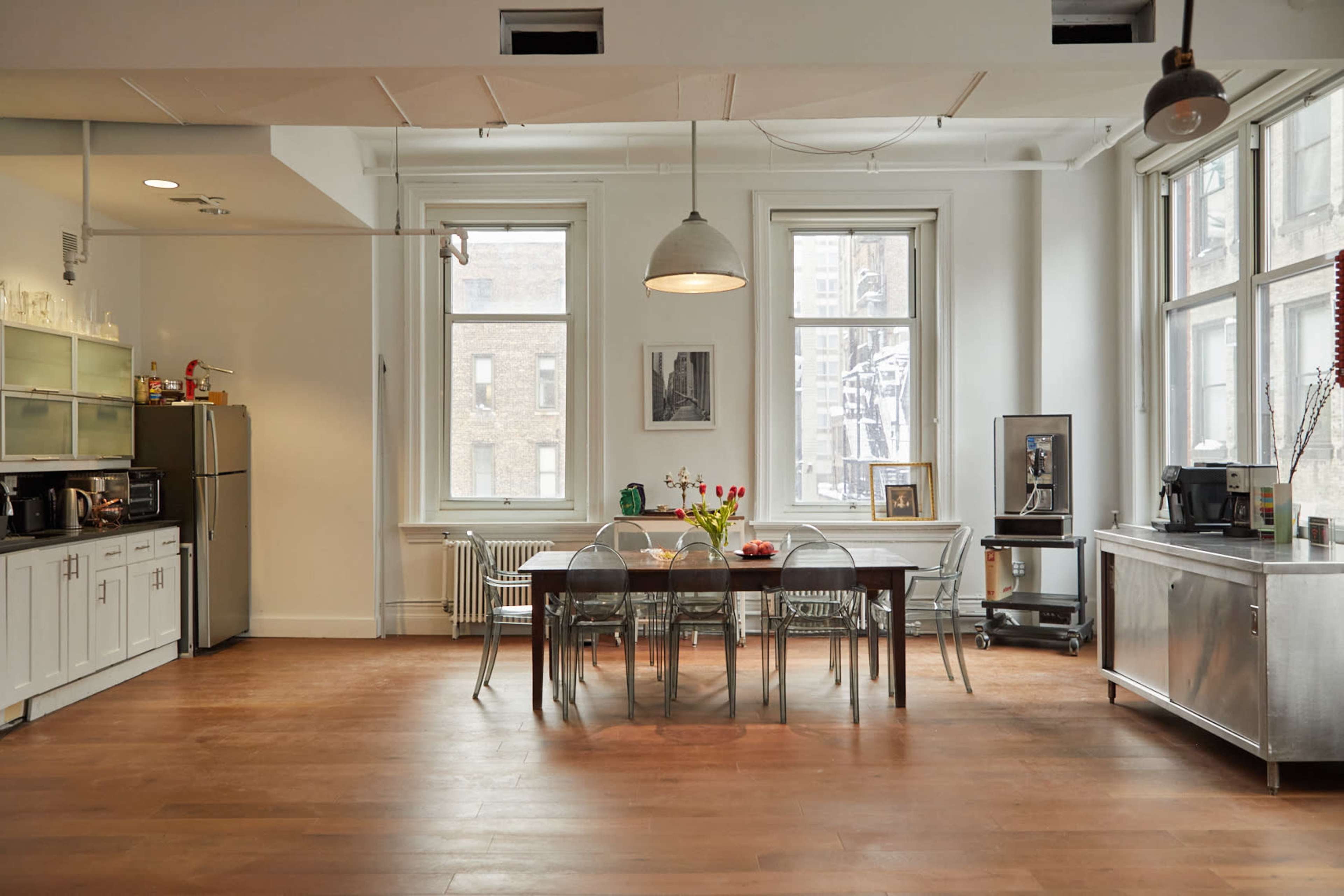 A bright kitchen and dining area features a long wooden table surrounded by clear chairs, with large windows allowing natural light to fill the space.