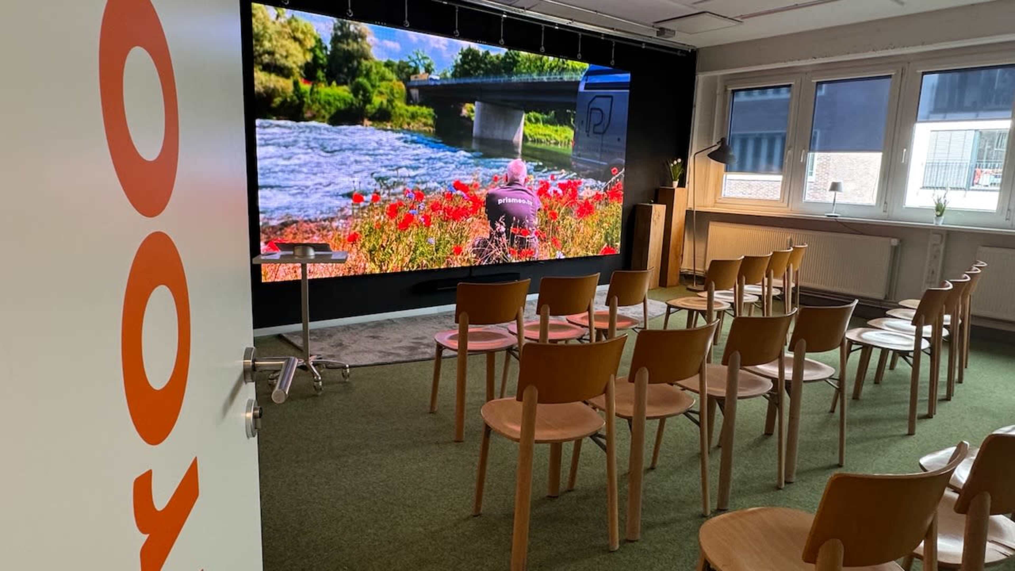 A modern room features a large screen displaying a landscape with a river and flowers, surrounded by rows of empty wooden chairs.