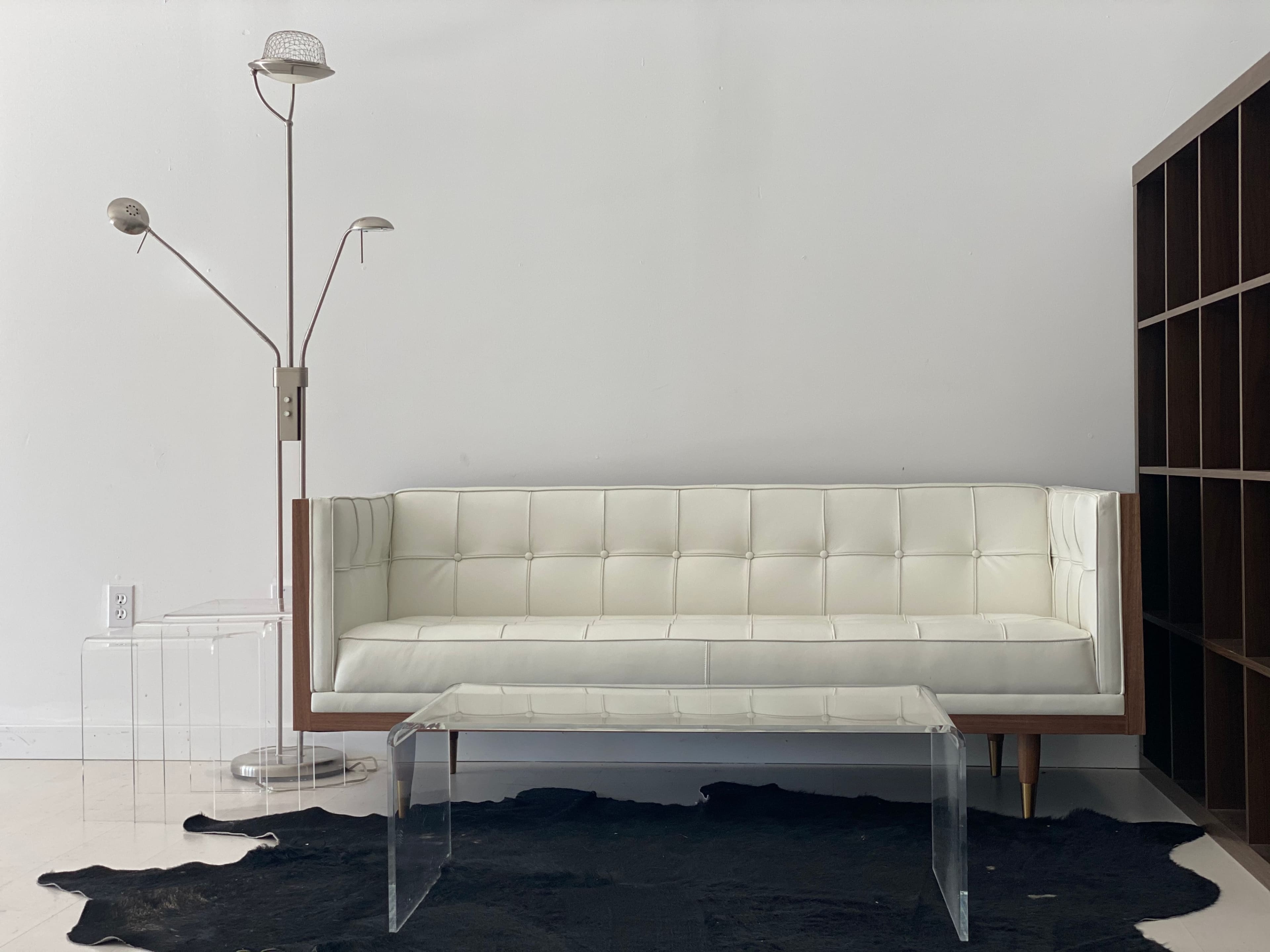 A white tufted sofa is positioned next to a floor lamp and a black cowhide rug, with a clear acrylic coffee table in front and an open shelving unit nearby.