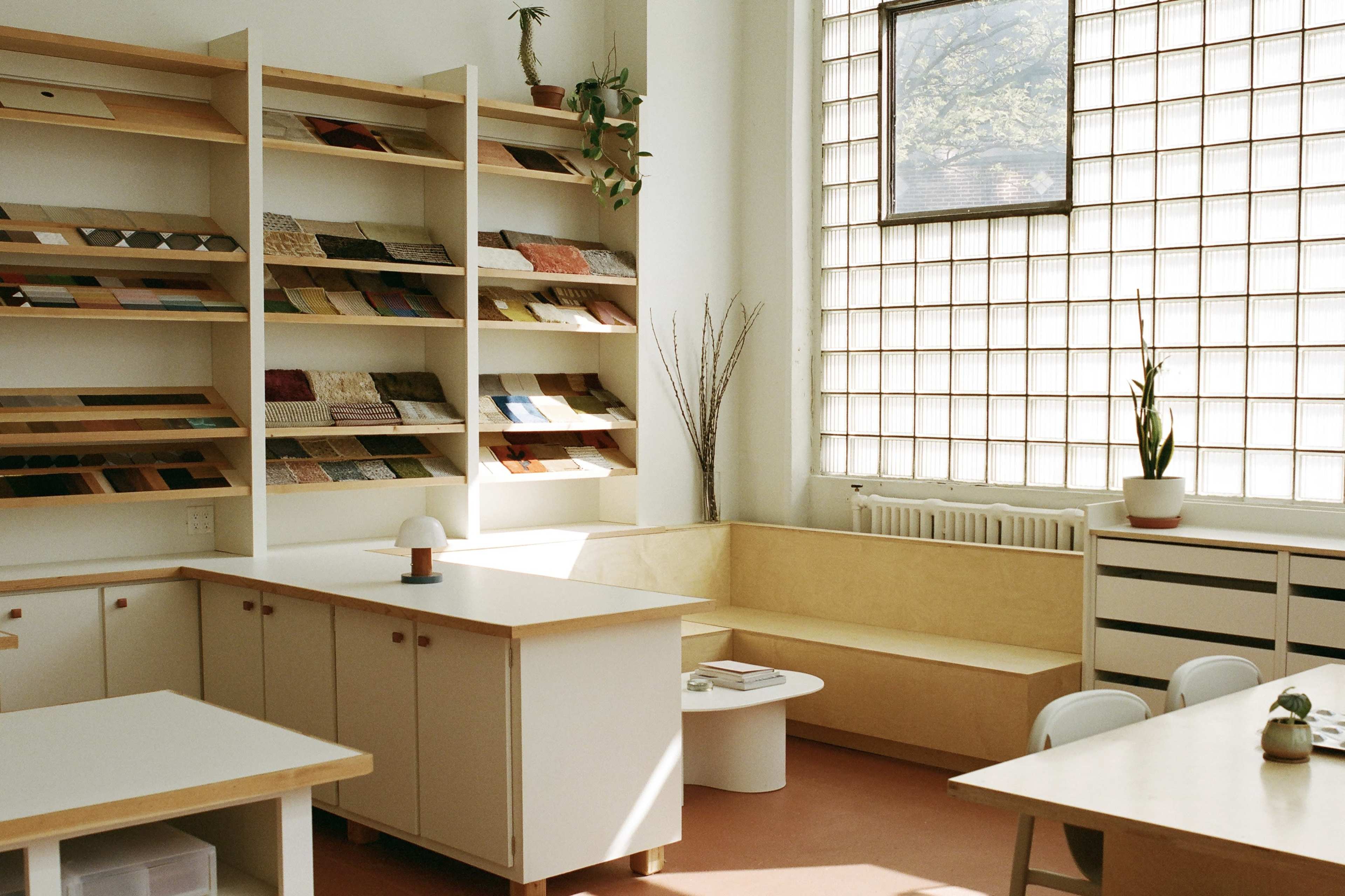 The image shows a bright, modern workspace featuring shelving filled with sample materials, a wooden bench, and a small round table.