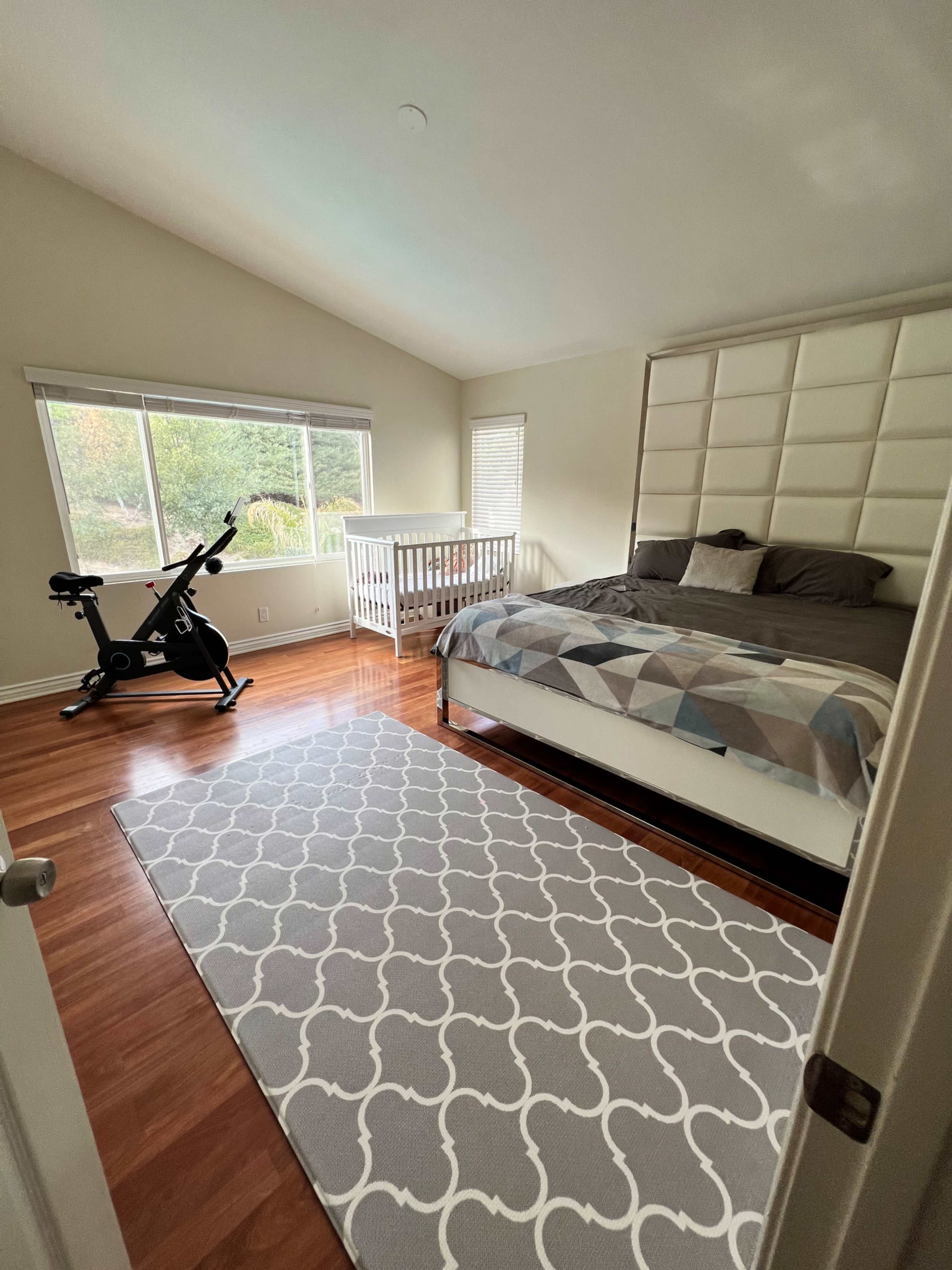A bedroom features a large bed with a patterned blanket, a crib by the window, a stationary bike, and a decorative rug on the wooden floor.