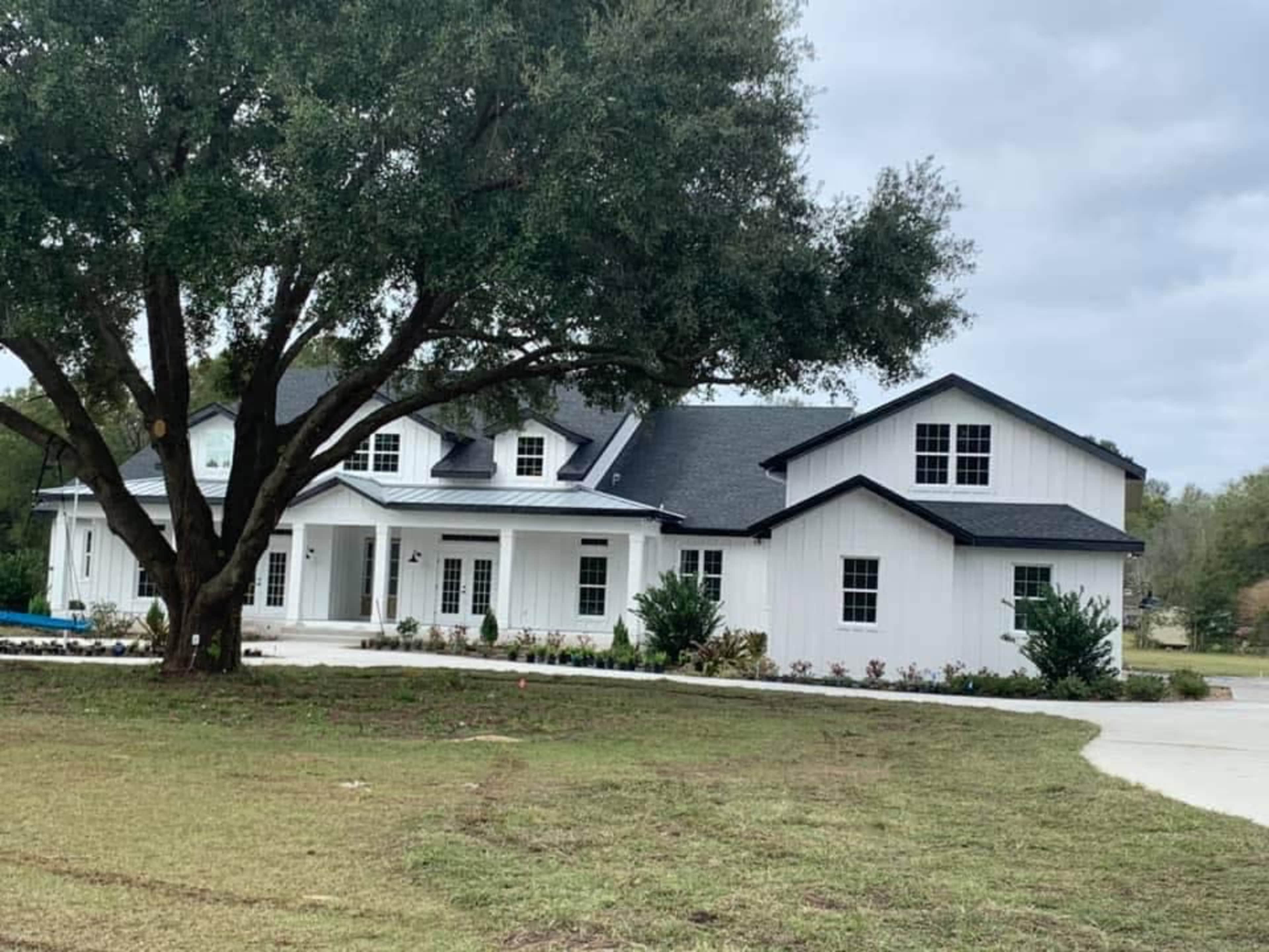 A large white house with a dark roof sits beside a grassy area and a large tree.