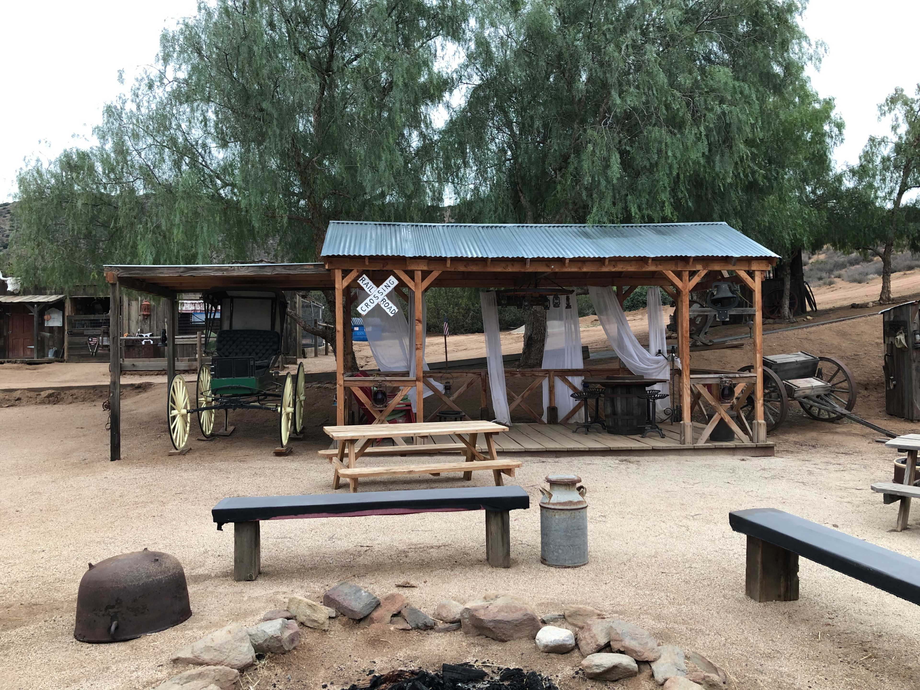 A rustic structure with a corrugated metal roof and wooden framework is situated next to a wagon under a covered area, surrounded by a sandy ground and sparse vegetation.