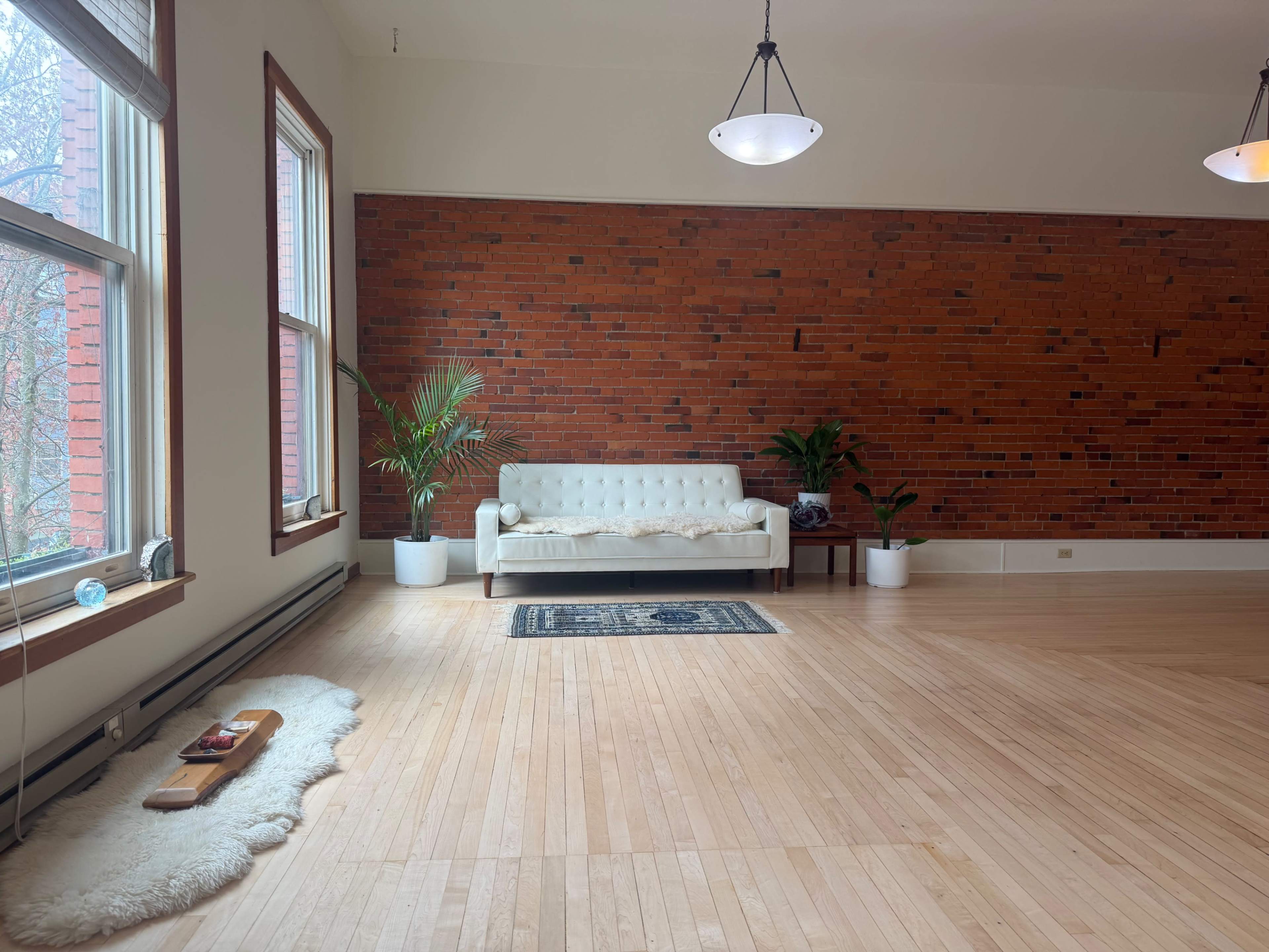 The image shows a spacious room with a brick accent wall, a white couch, plants, and wooden flooring.
