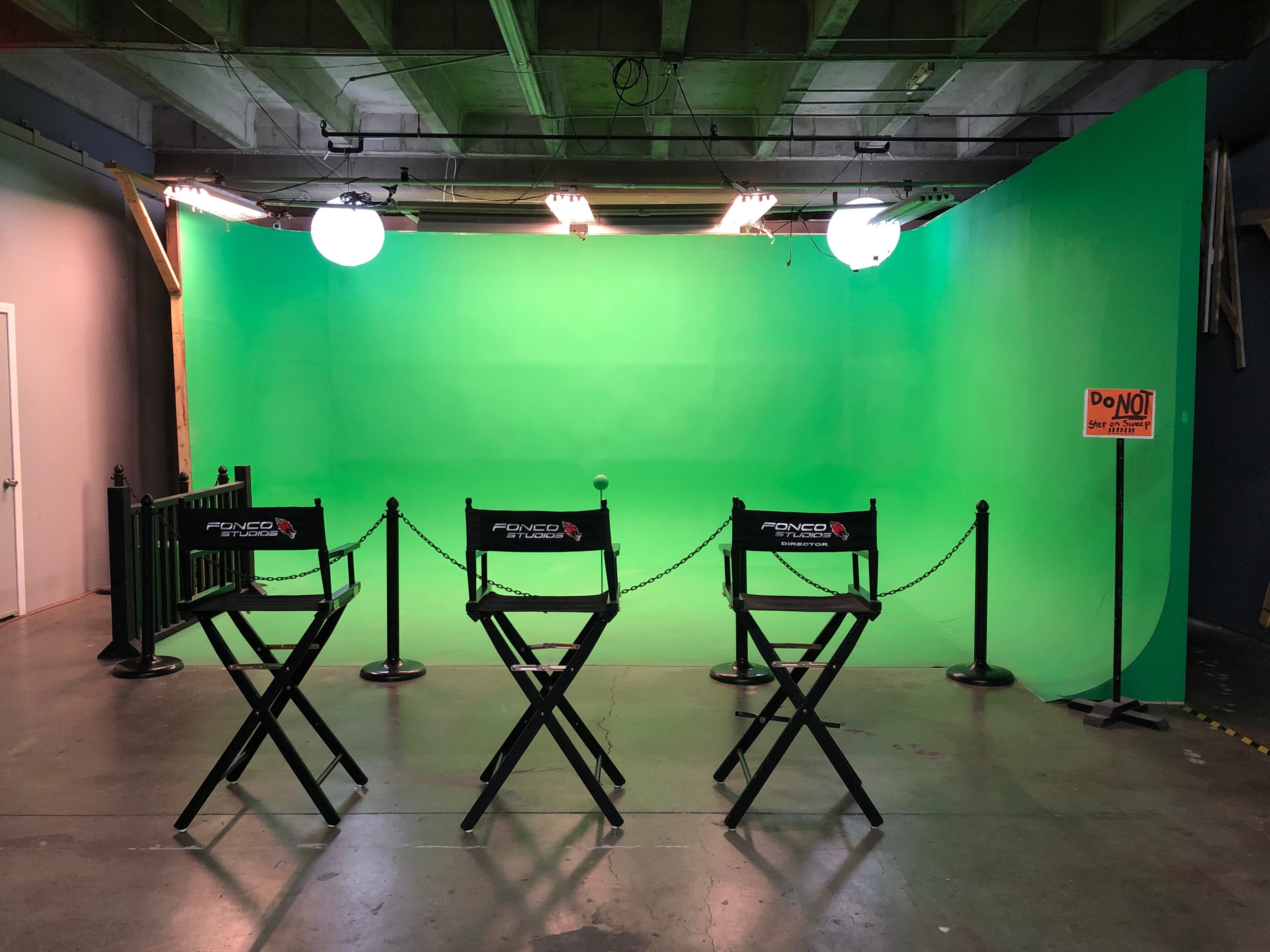 Three empty director's chairs in front of a green screen setup, with bright lights hanging above.
