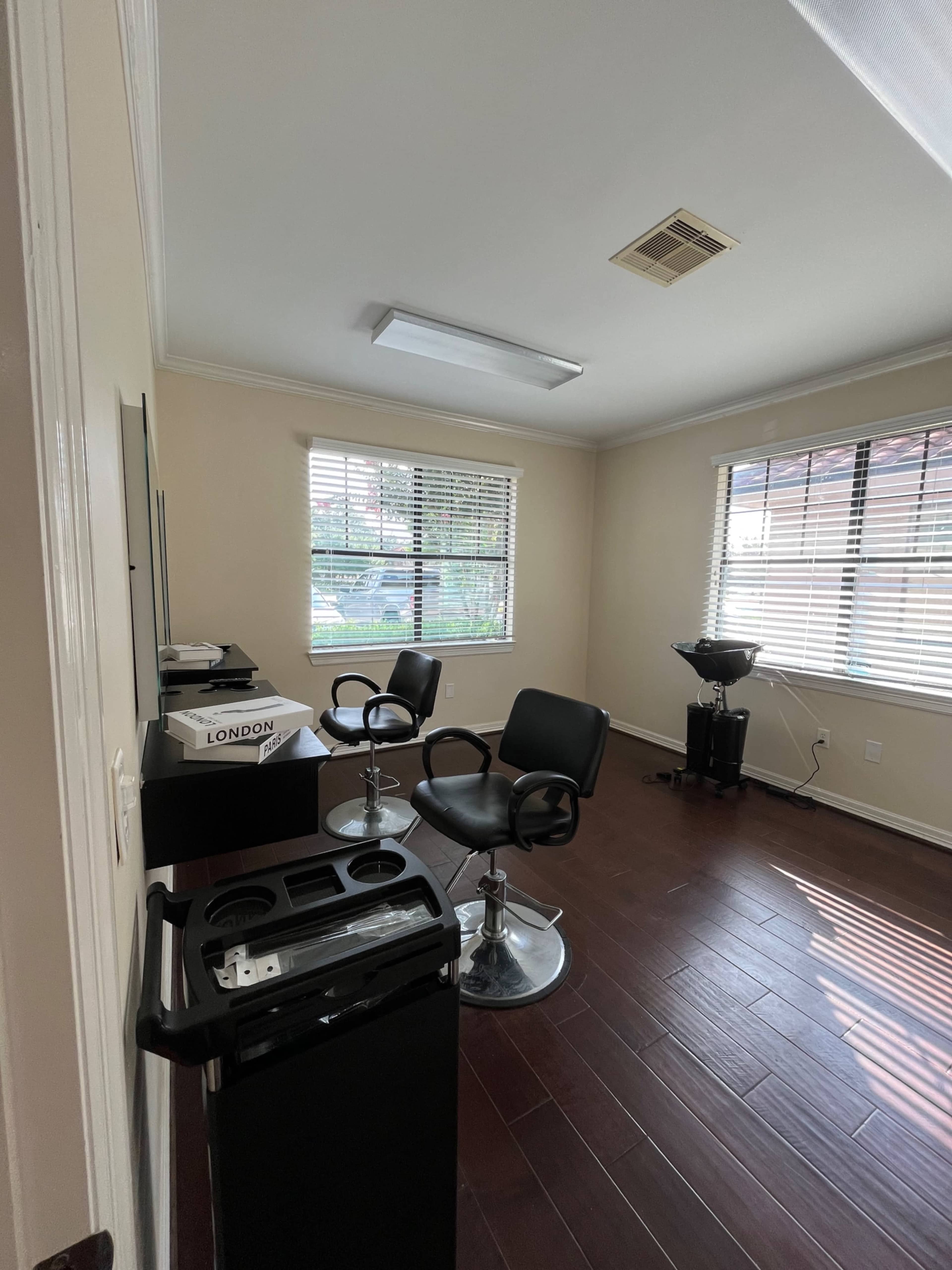 The image shows a sparsely furnished salon room with two black salon chairs, a hair wash station, and a reception area on one side.
