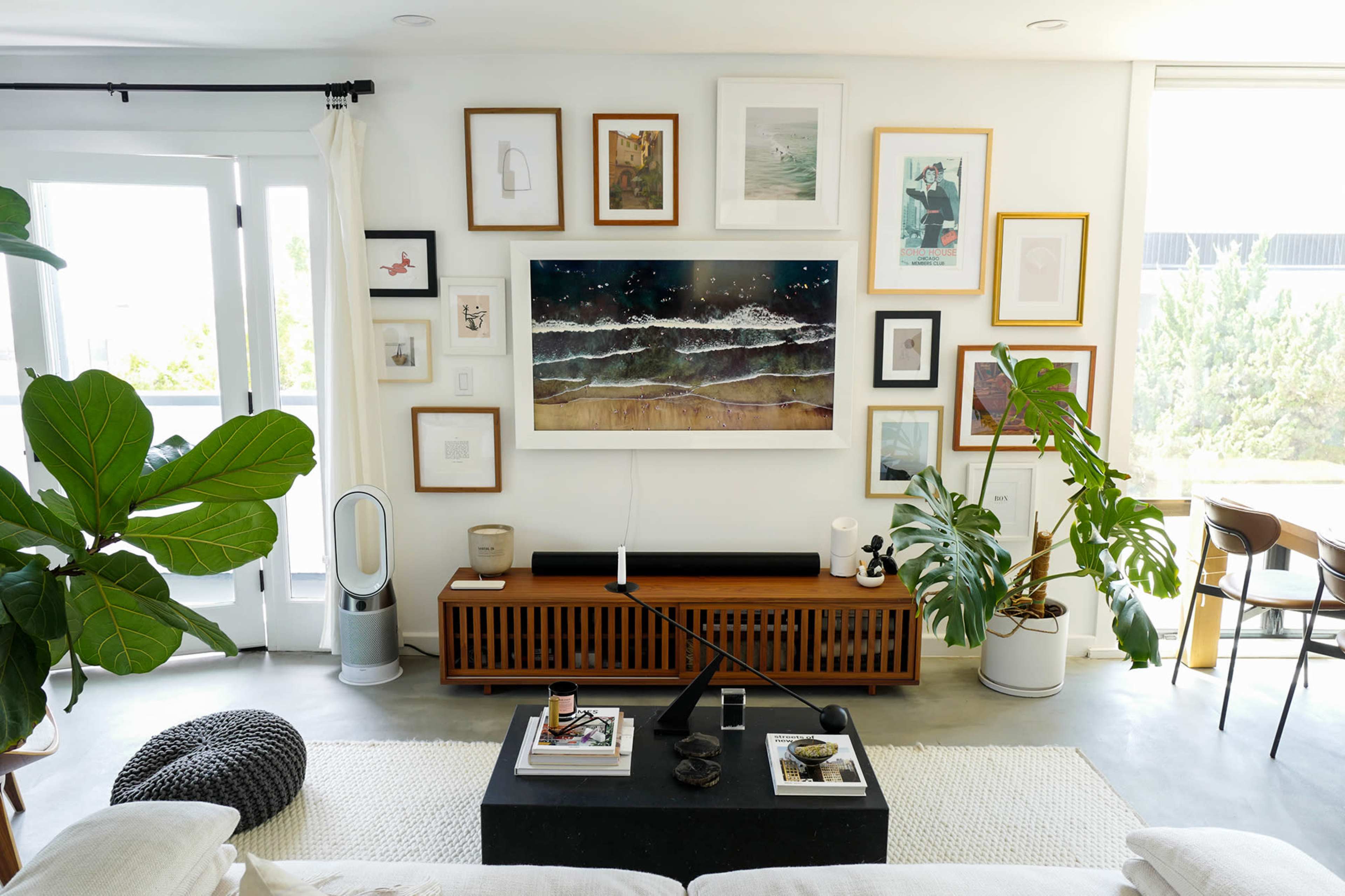 A modern living room features a wall adorned with various framed artworks, a television mounted above a wooden media console, and two large indoor plants beside a coffee table.