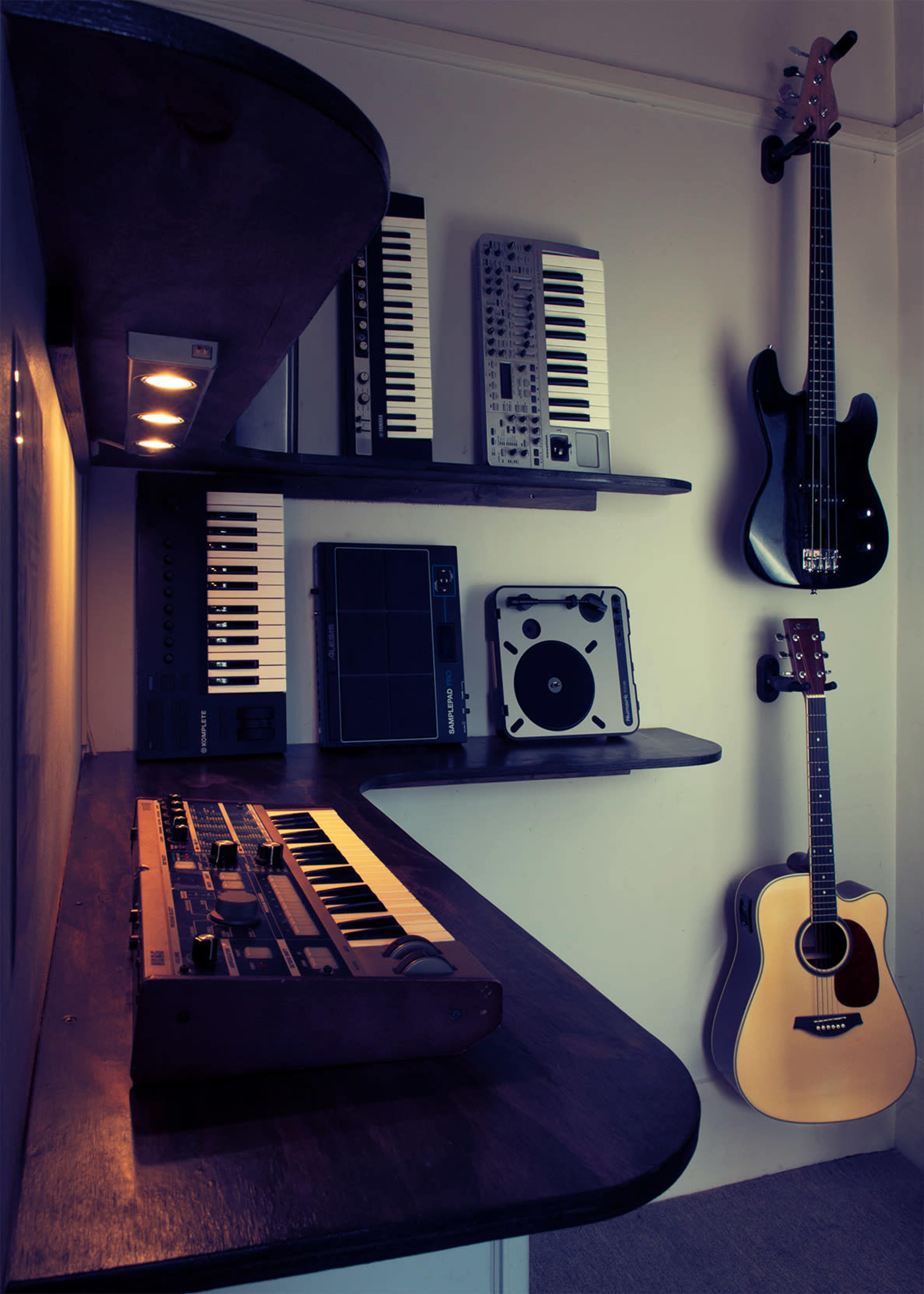 The image shows a collection of musical instruments and equipment displayed on shelves, including keyboards, a drum pad, and an acoustic guitar, mounted on a wall.