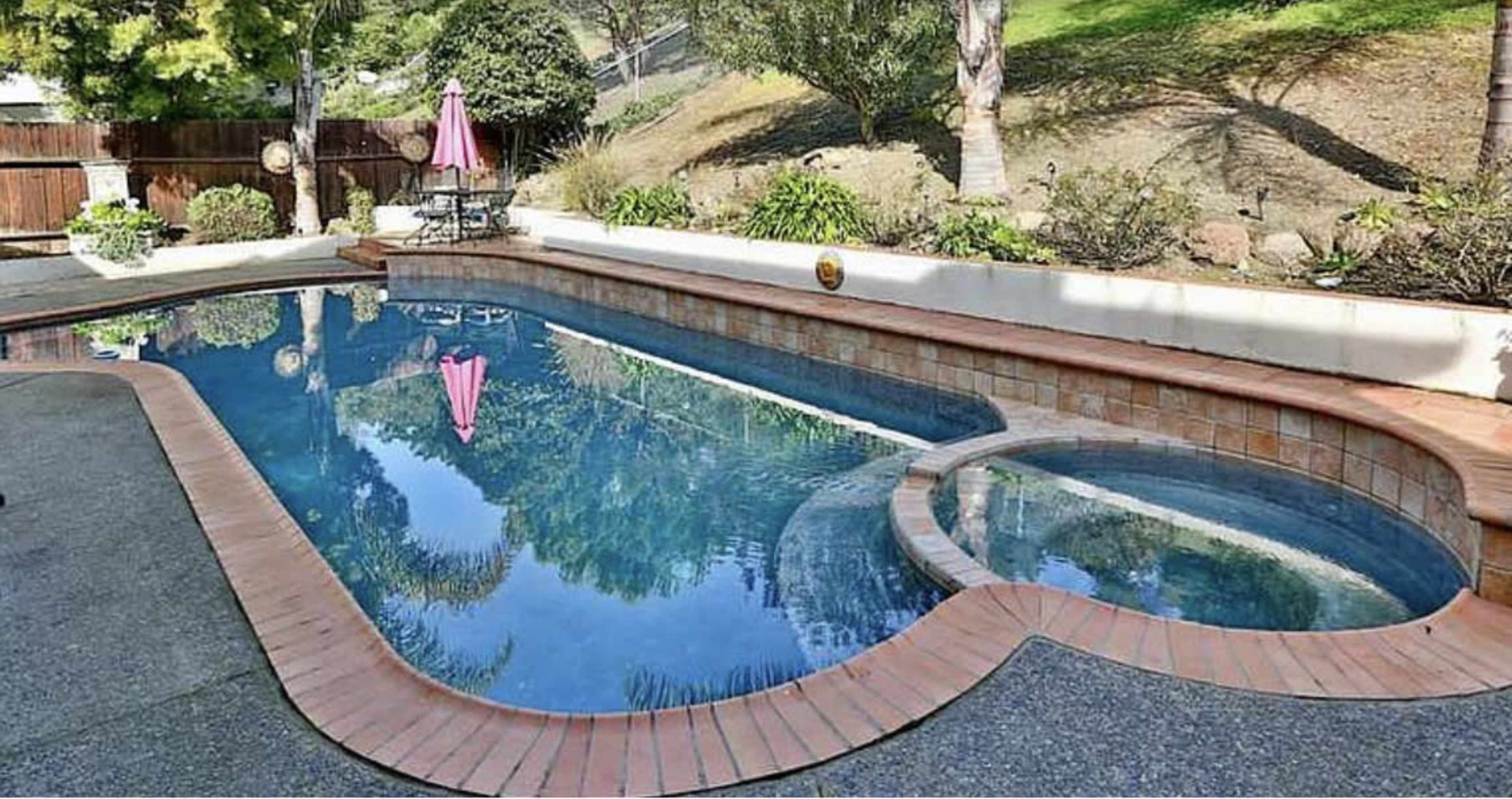 A backyard swimming pool with a heart-shaped design, bordered by a brick walkway and surrounded by greenery.