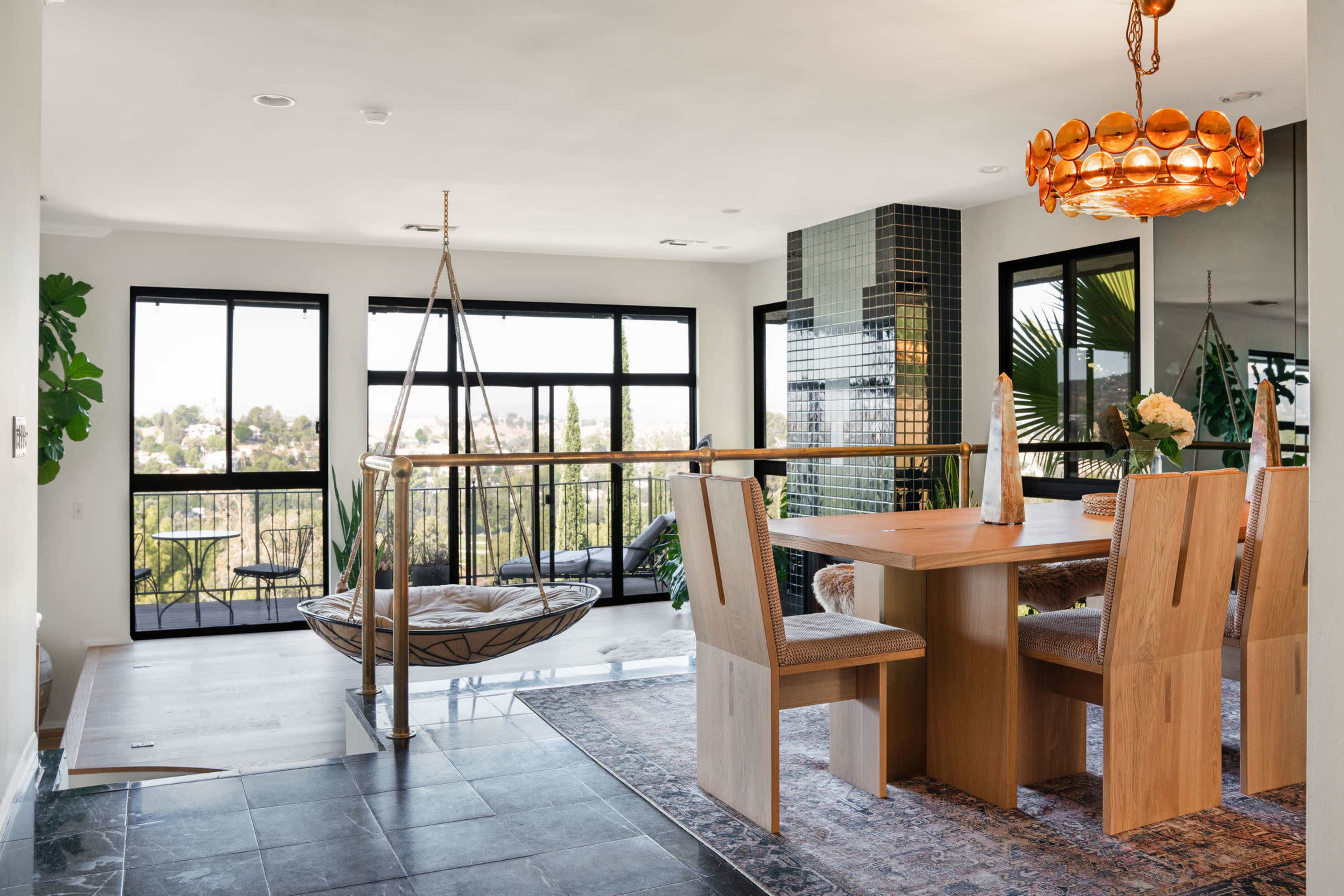A modern dining area featuring a wooden table surrounded by light-colored chairs, a hanging chair, and large windows offering a view of the outdoors.