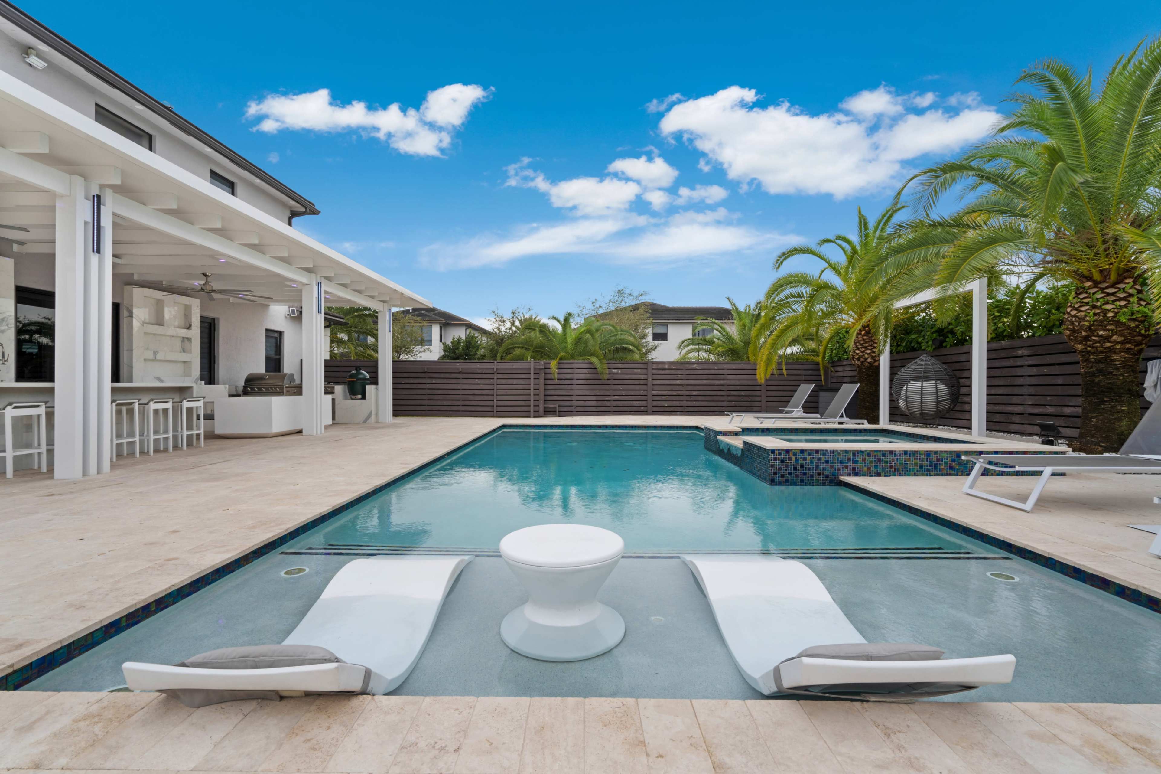 A modern backyard features a swimming pool surrounded by palm trees, lounge chairs, and a covered outdoor seating area.
