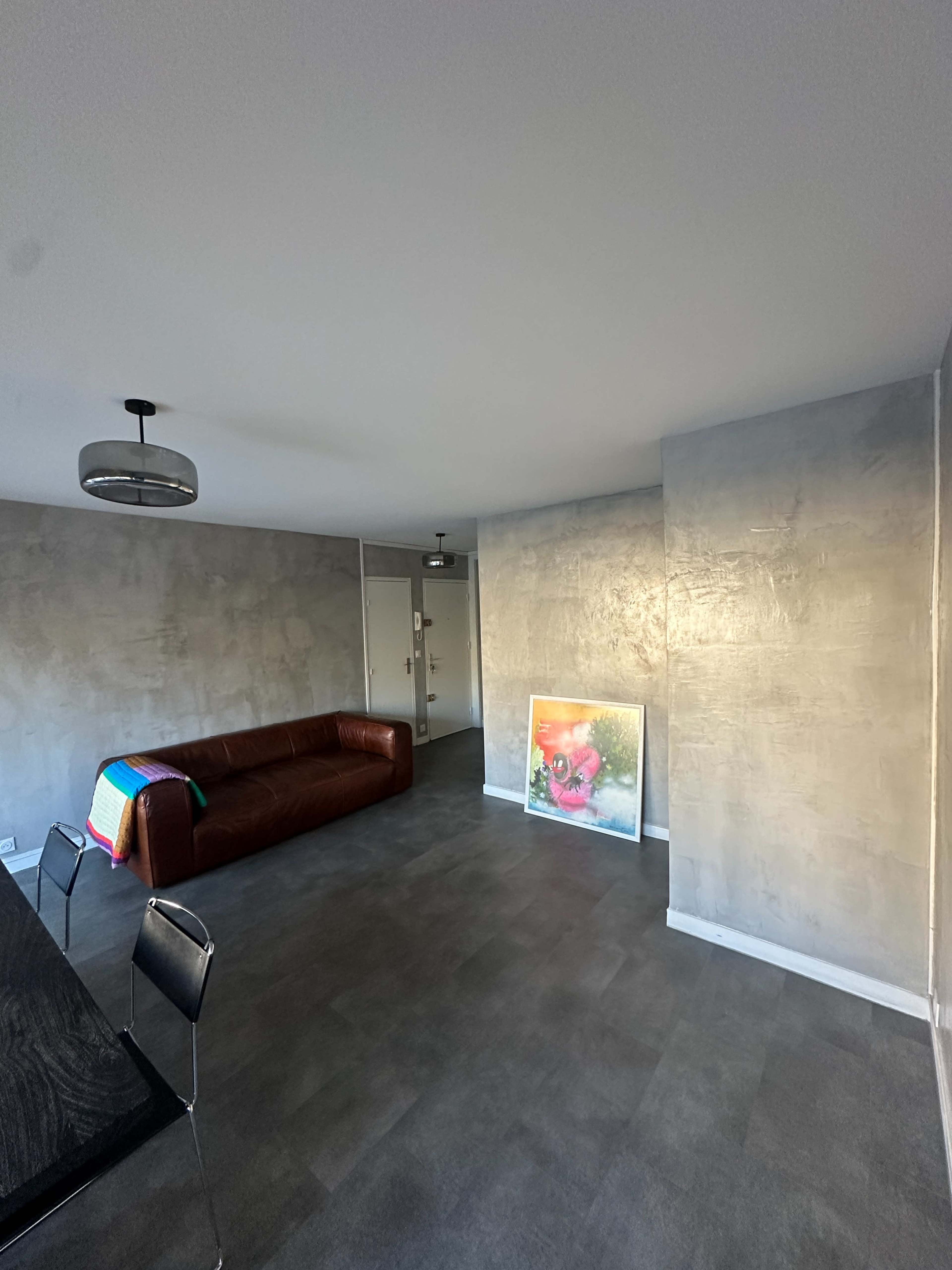 The image shows a spacious living room with a brown leather sofa, a dining table with black chairs, and a colorful painting leaning against the wall.