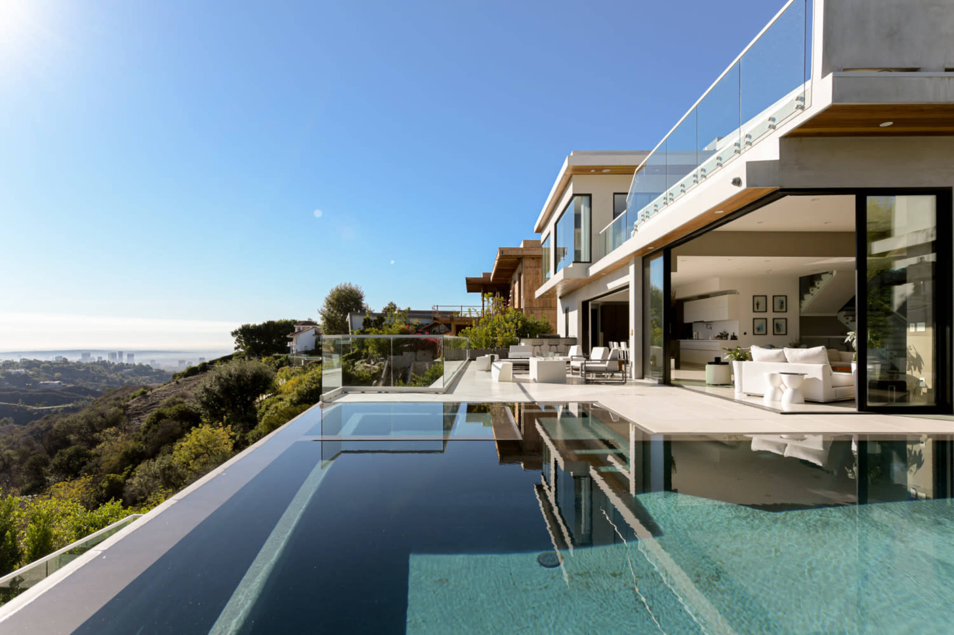 A modern villa with large glass walls overlooks a swimming pool and a panoramic view of the hills and city below.