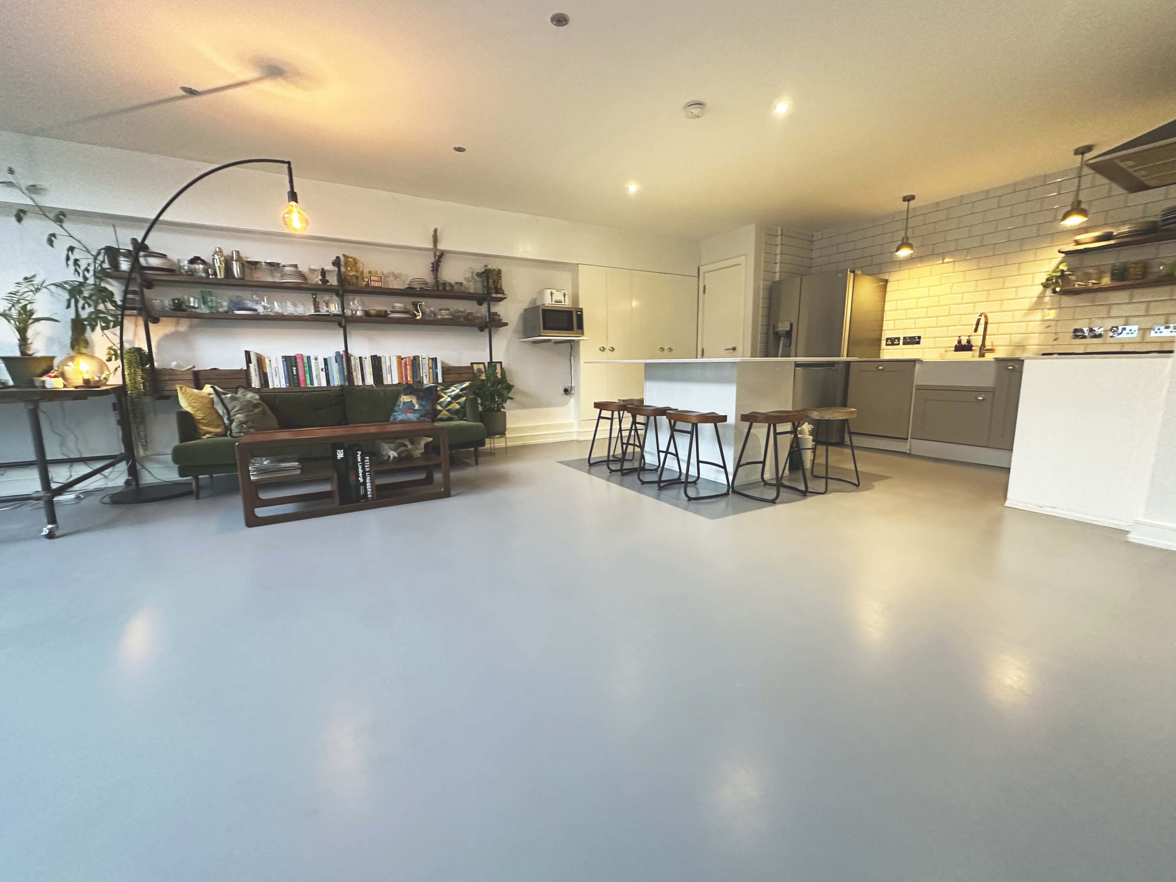 A modern open-concept living space featuring a green sofa and a wooden coffee table on the left, with a kitchen area equipped with stainless steel appliances and a bar with stools on the right.