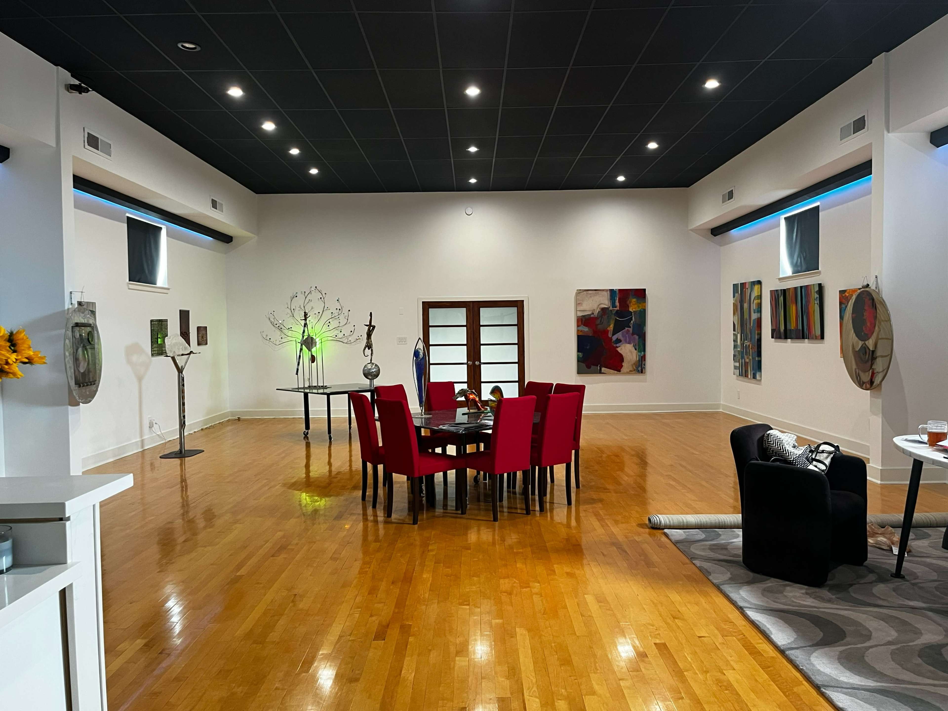 A spacious room features a round table surrounded by red chairs, with various artworks and decorative objects placed throughout.