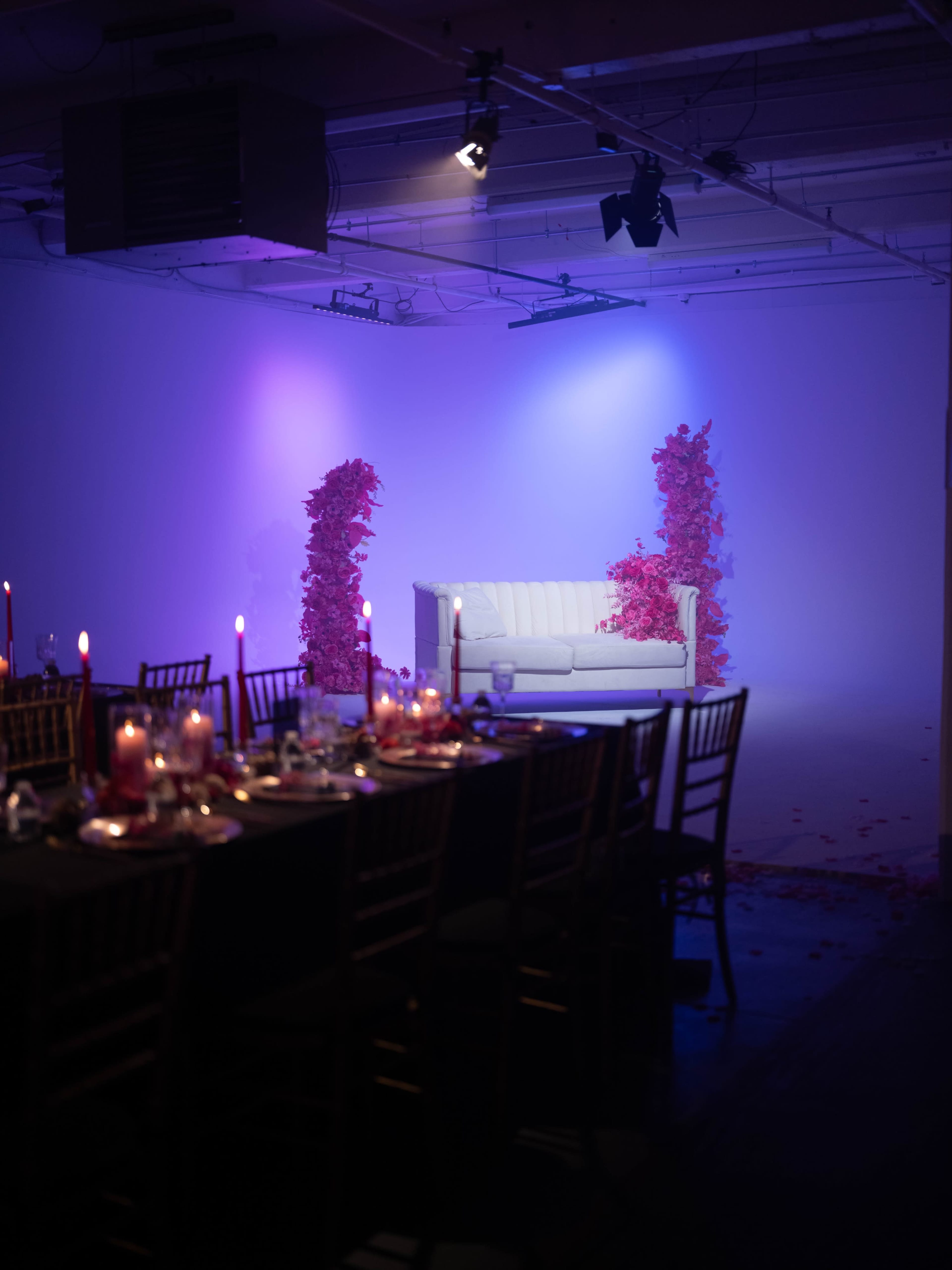 A white sofa is centered in a dimly lit room, flanked by tall arrangements of pink flowers, with a long dining table set in the foreground.
