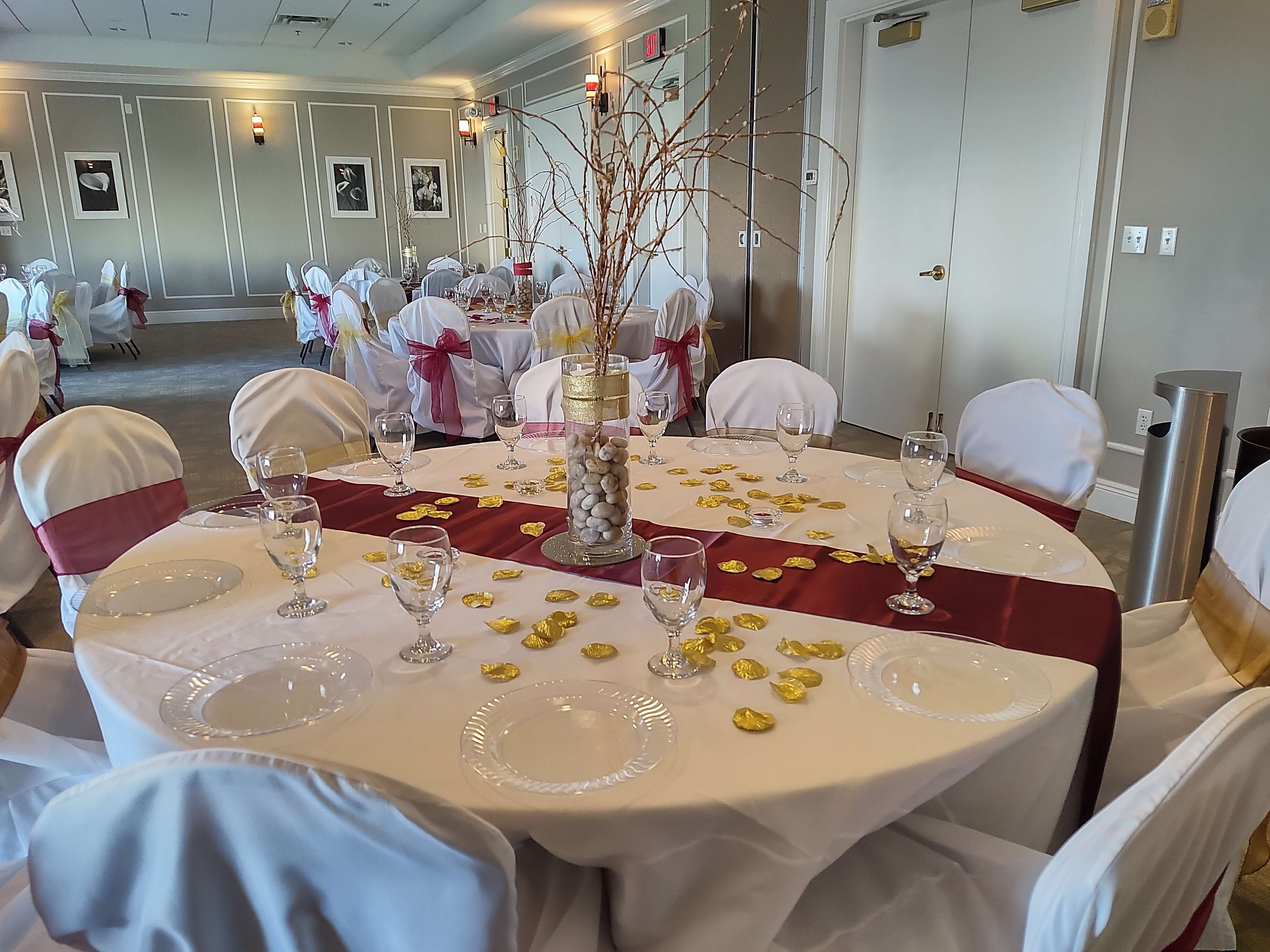 A dining area is set with a table adorned with a white tablecloth, red runner, glassware, and decorative items, surrounded by chairs covered in white and red fabric.