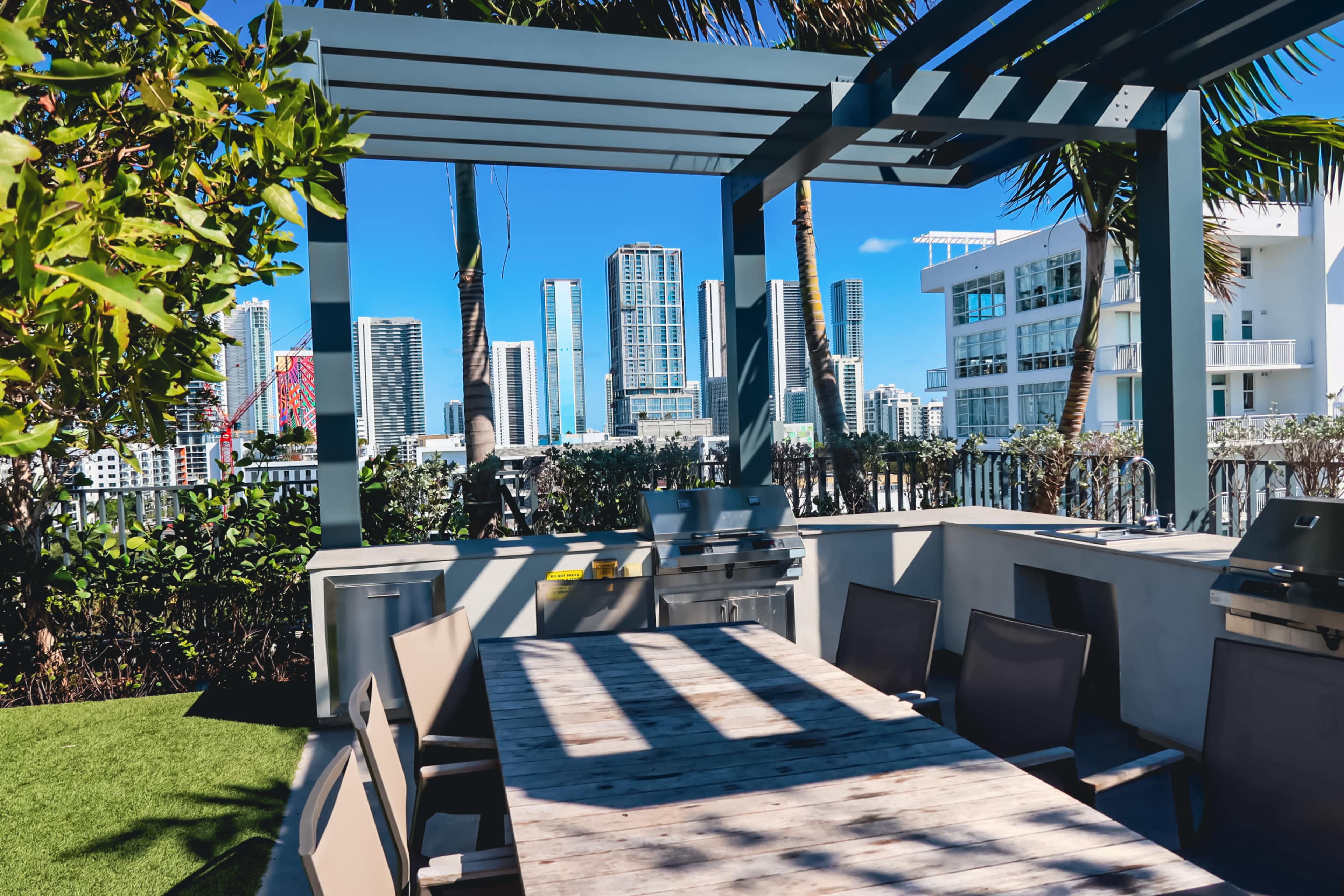 The image shows an outdoor dining area with a wooden table and a grill, set against a backdrop of tall city buildings under a clear blue sky.