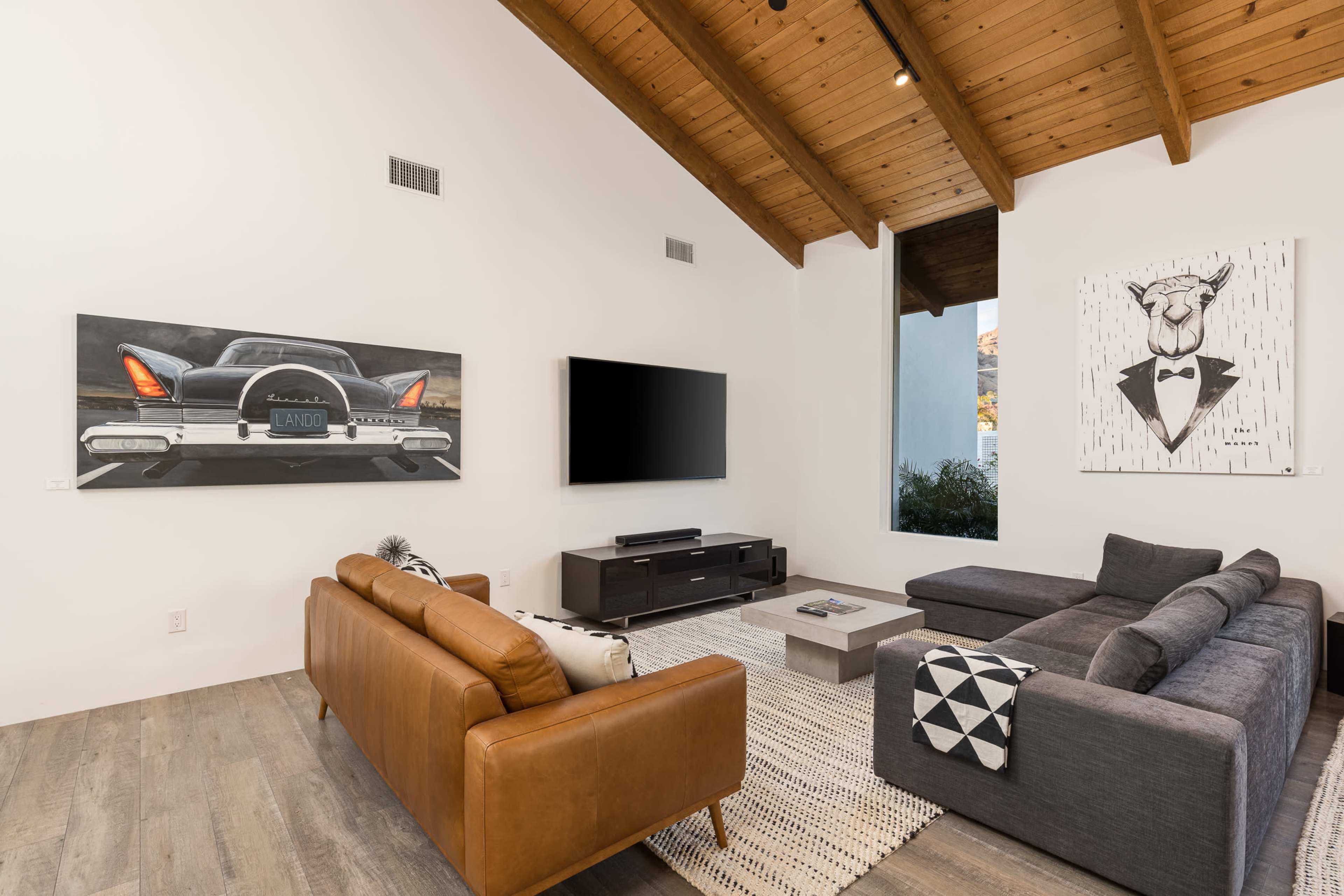 A modern living room features a large sectional sofa, a leather armchair, a flat-screen TV, and artwork on the walls, with vaulted wooden ceilings and natural light coming through a window.