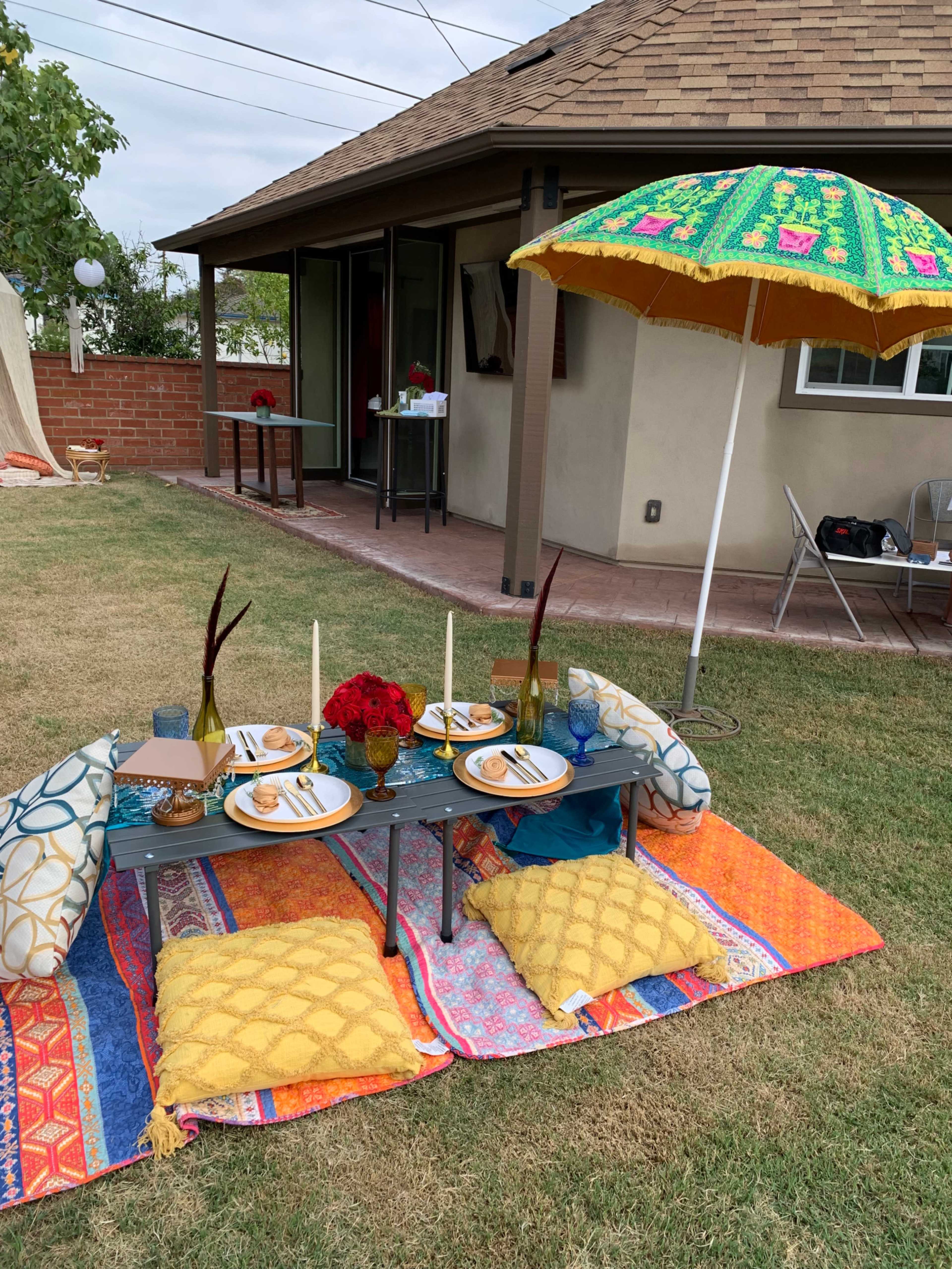 A colorful outdoor dining setup features a low table with plates, glasses, and flowers, surrounded by cushions on a patterned blanket under a decorative umbrella.