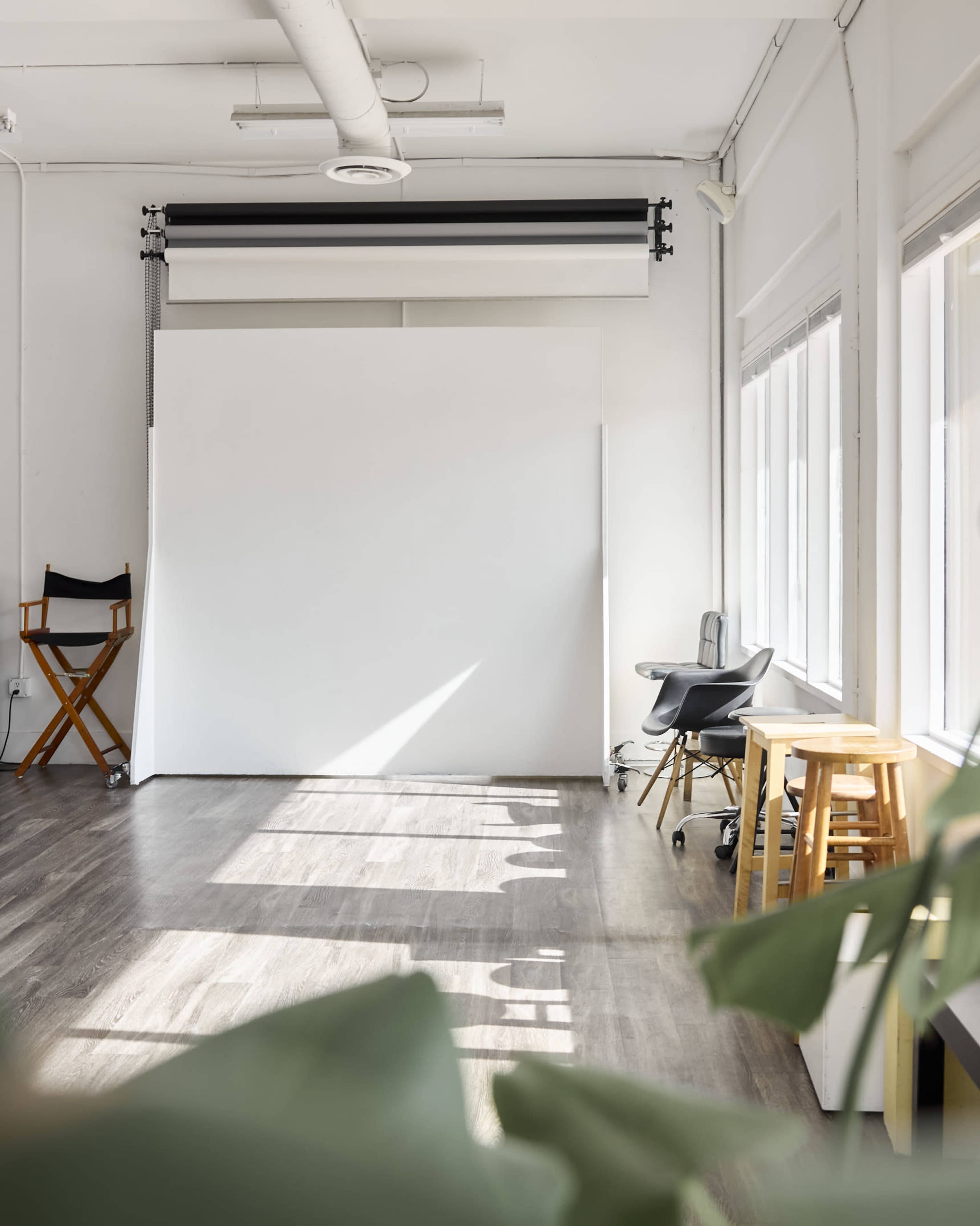 A bright, minimalist studio space features a large white backdrop, wooden flooring, and a few pieces of simple furniture, including a director's chair and a small stool, with natural light streaming through the windows.
