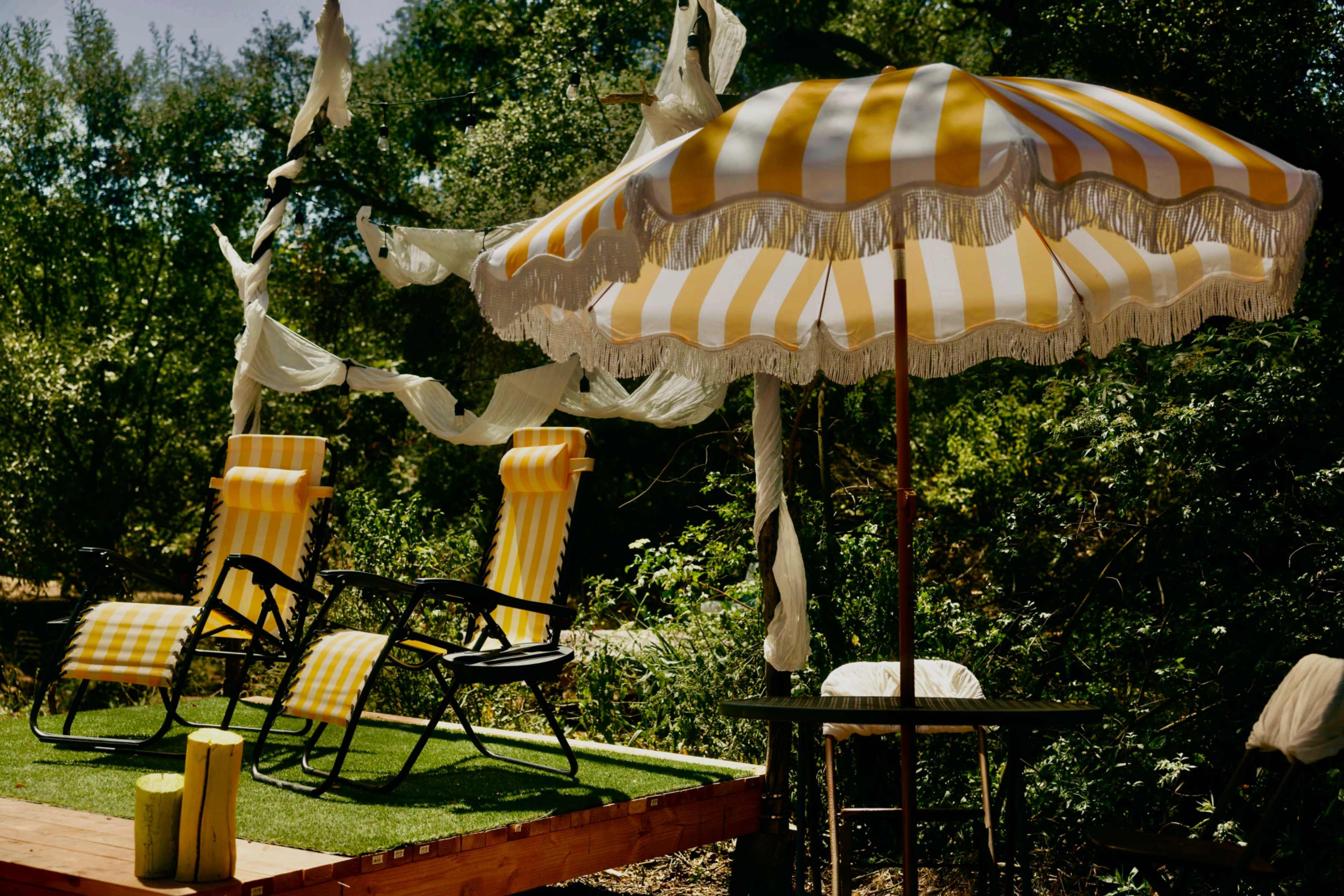 The image shows a small outdoor relaxation area with two yellow striped lounge chairs, a matching umbrella, and greenery in the background.
