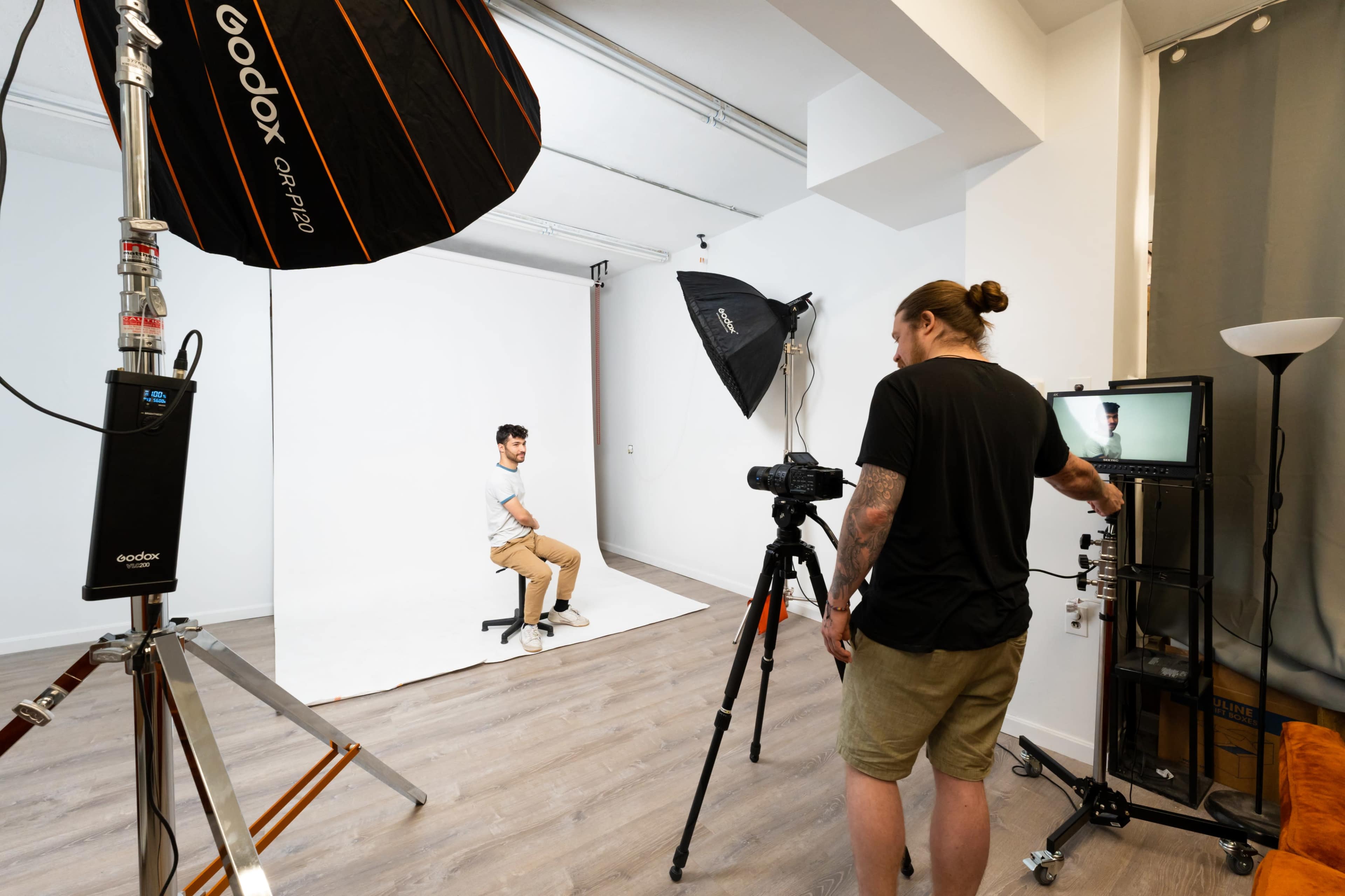 A photographer sets up a studio scene with lighting equipment and a camera while a model poses on a stool in front of a plain white backdrop.