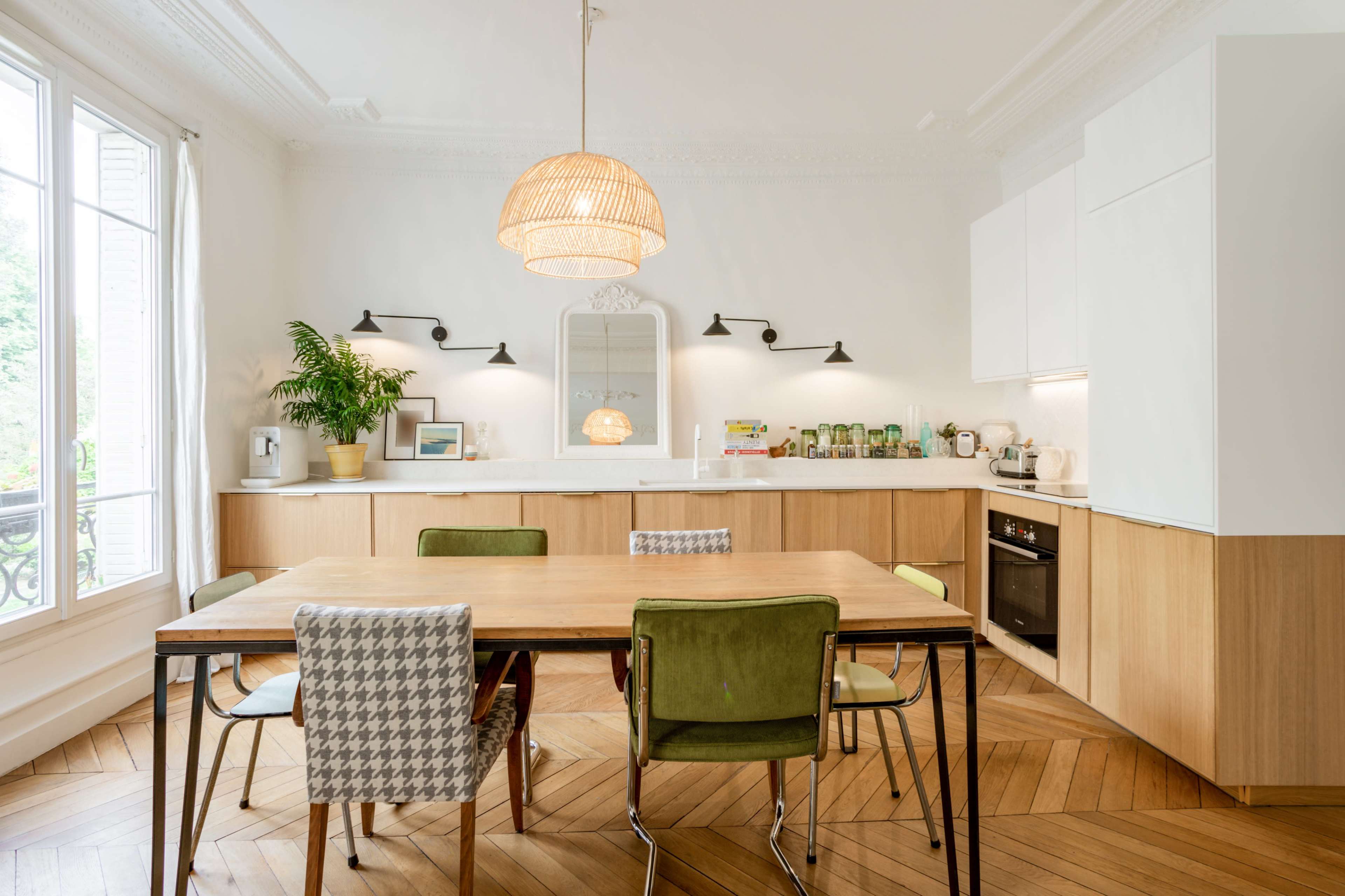 A modern kitchen features a wooden dining table surrounded by various chairs, with white cabinets and a large window allowing natural light to enter.