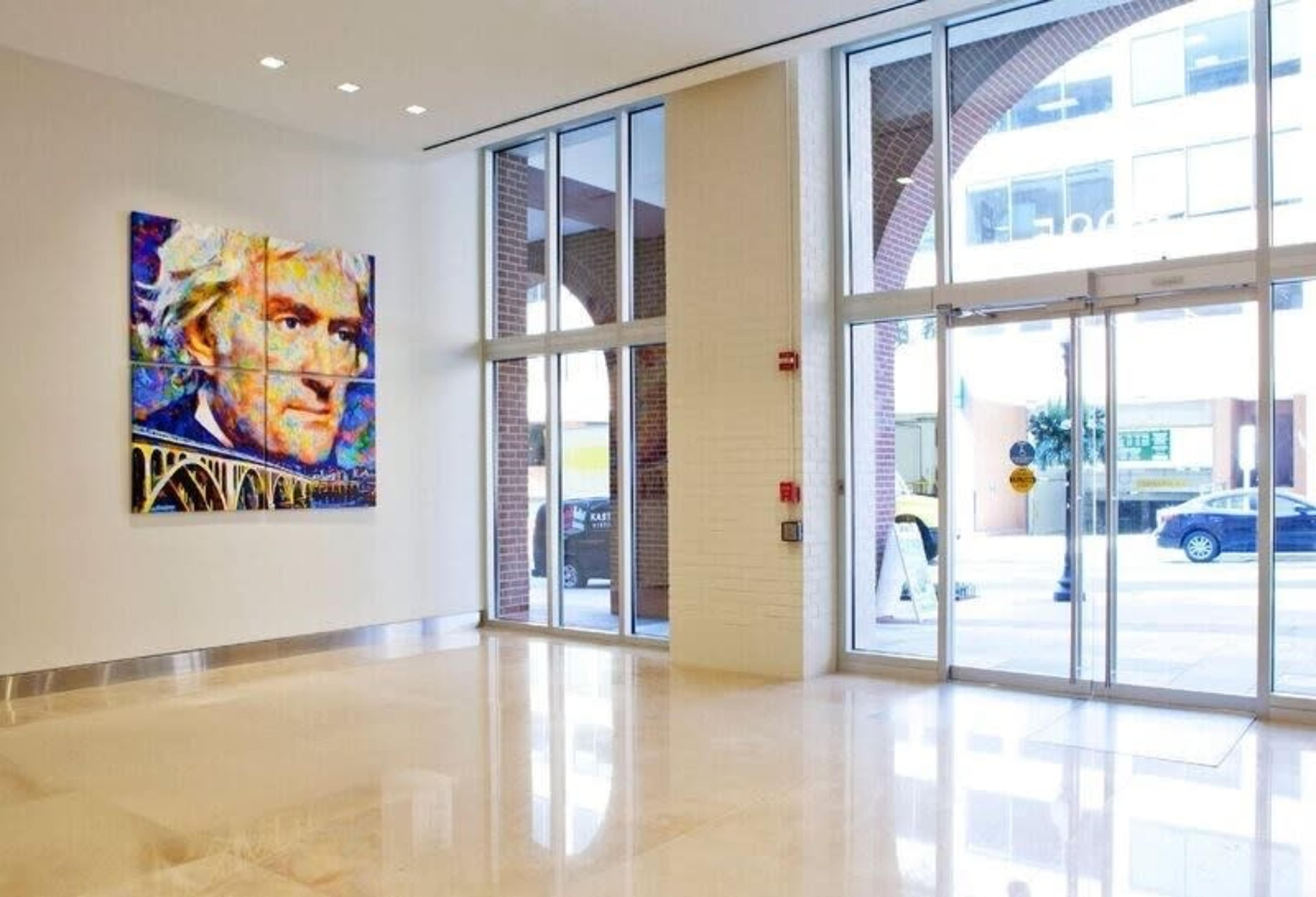 The image shows a spacious lobby with a large colorful portrait of a man on the wall, large windows allowing natural light, and a glass door leading to the street outside.