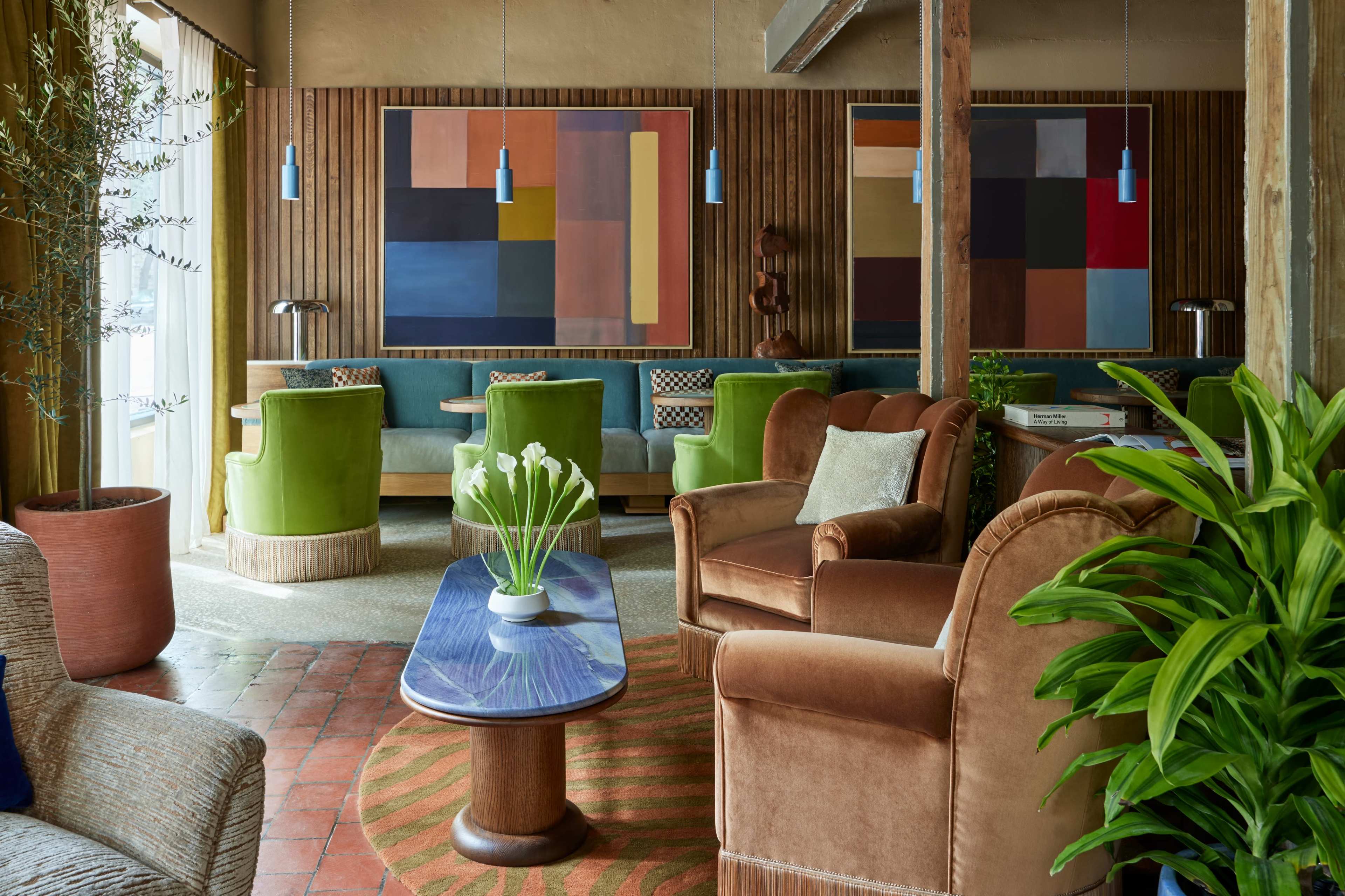 The image features a stylish lounge area with colorful abstract art, green and brown upholstered seating, and a wooden table surrounded by potted plants.