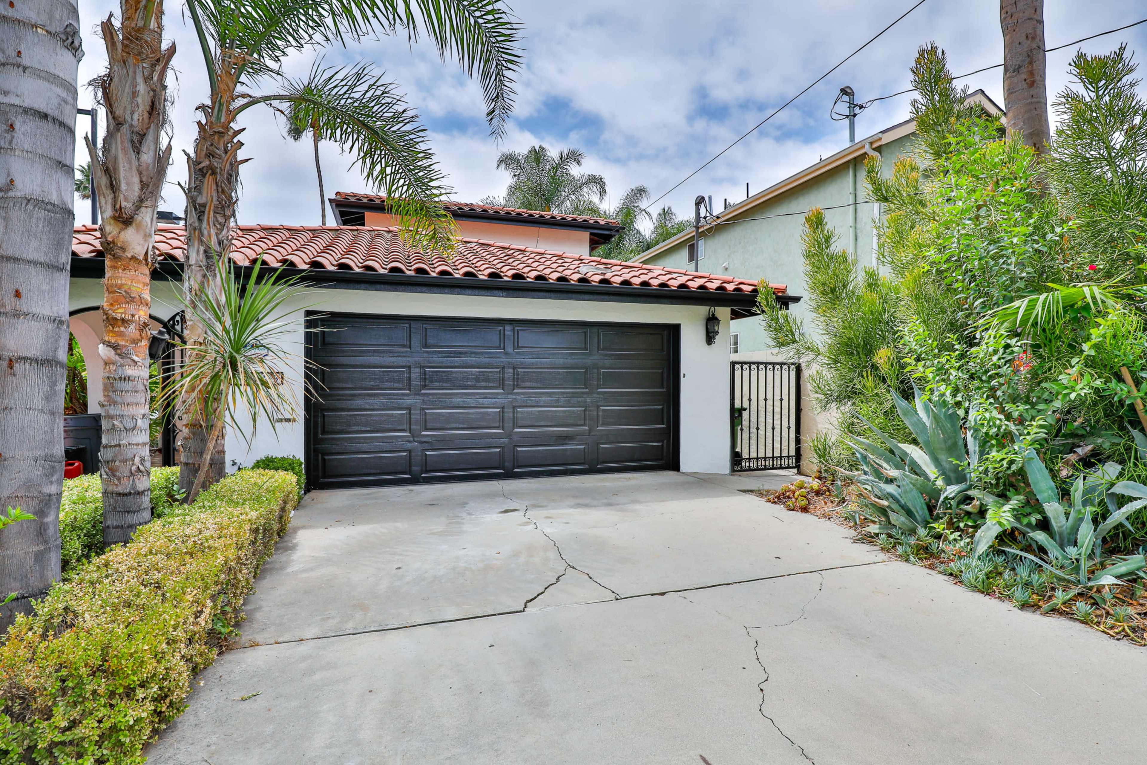 A driveway leads to a house featuring a dark garage door, surrounded by palm trees and various lush plants.