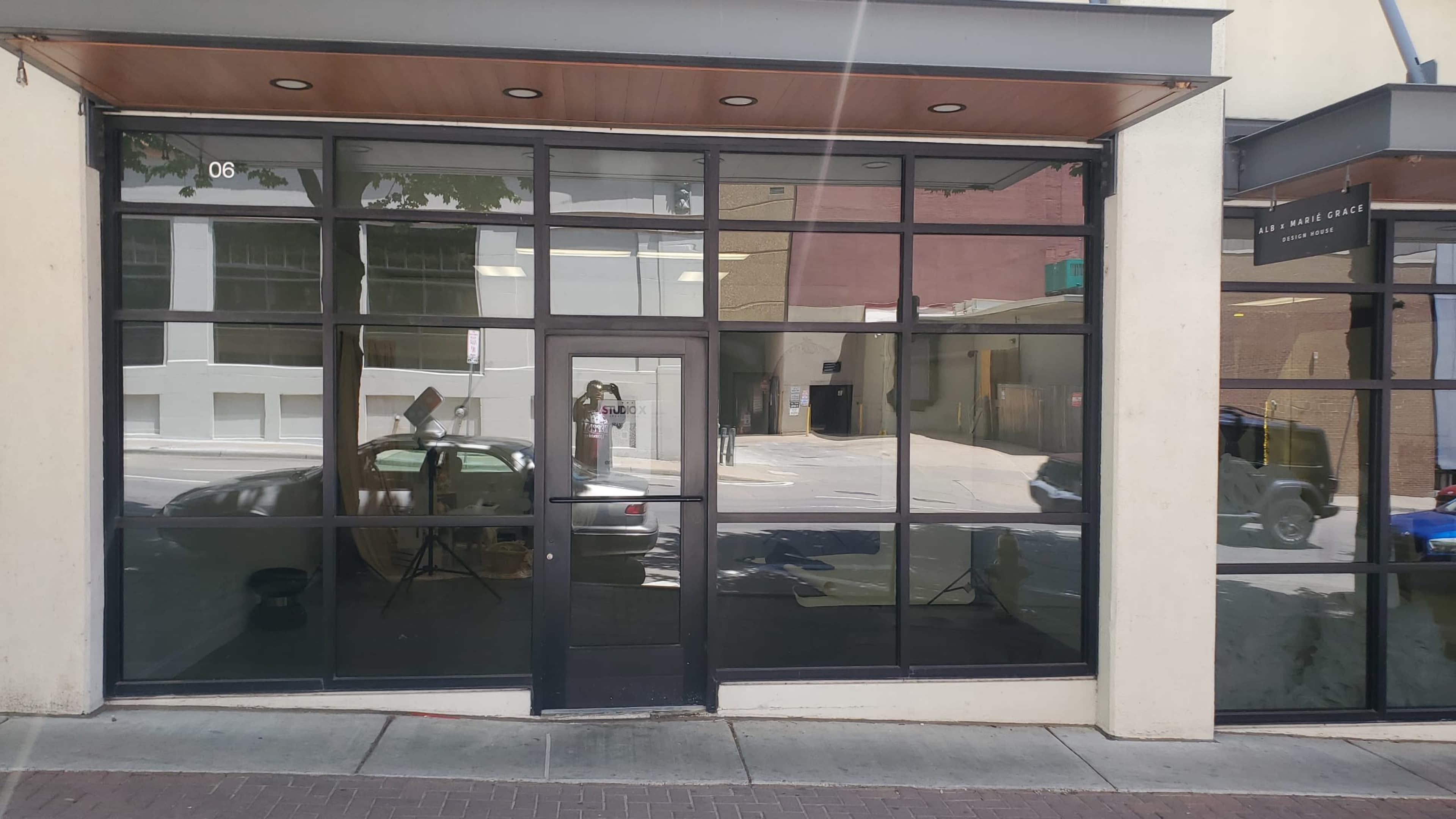 The image shows a commercial storefront with large glass windows and a black entrance door, reflecting a nearby street and vehicles.