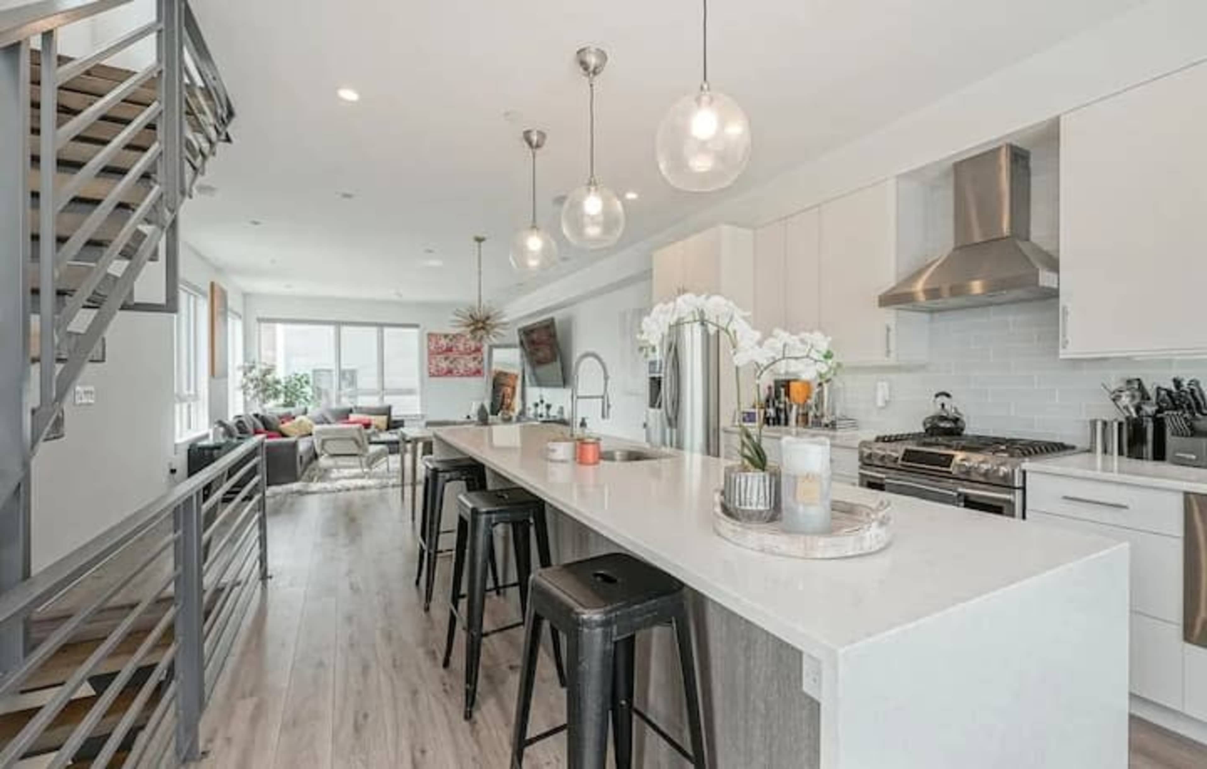 A modern kitchen and living area with a long white island, bar stools, stainless steel appliances, and large windows.