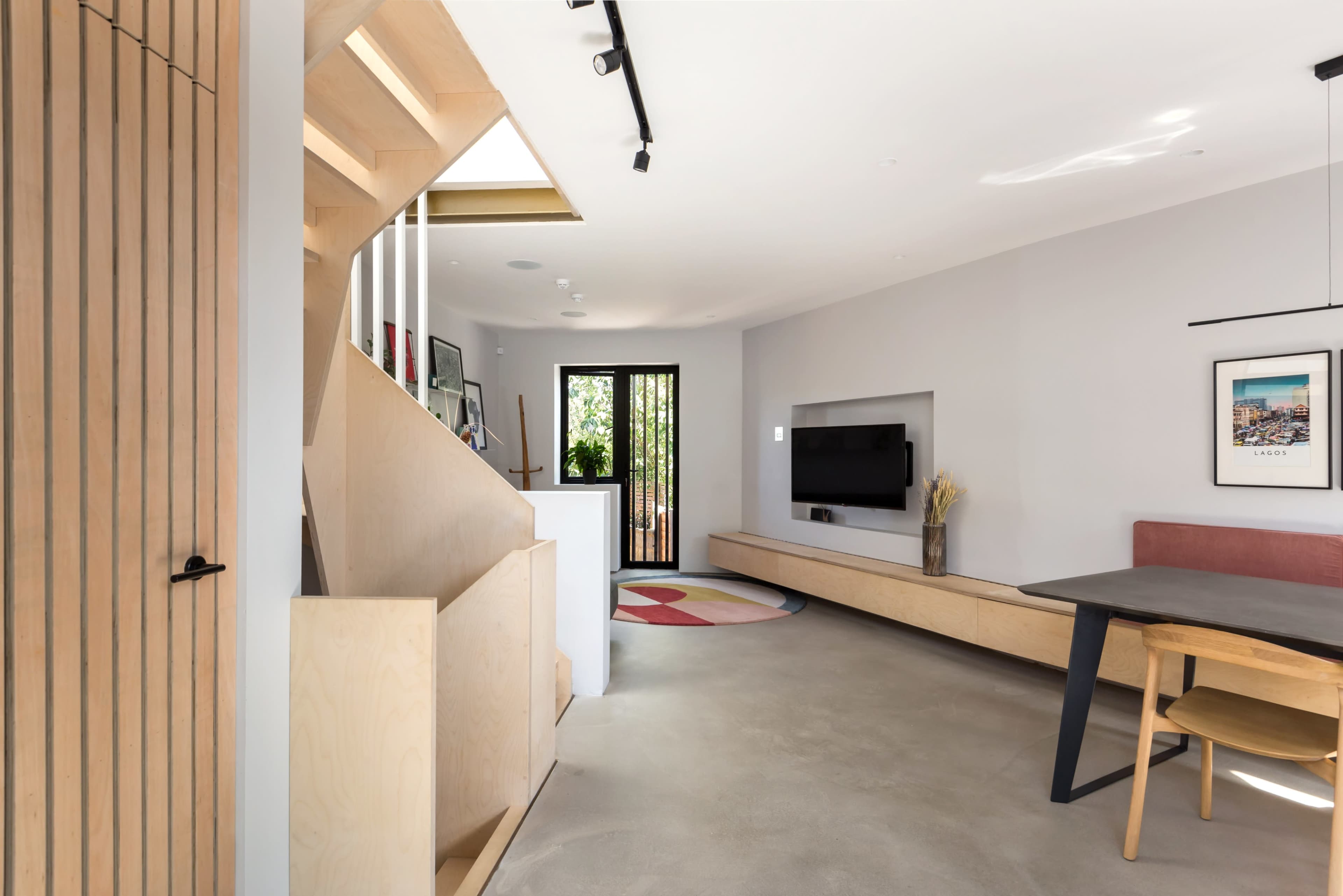 A modern living space with a staircase, a wall-mounted TV, a minimalist wooden shelf, and a dining area with a dark table and light-colored chairs.