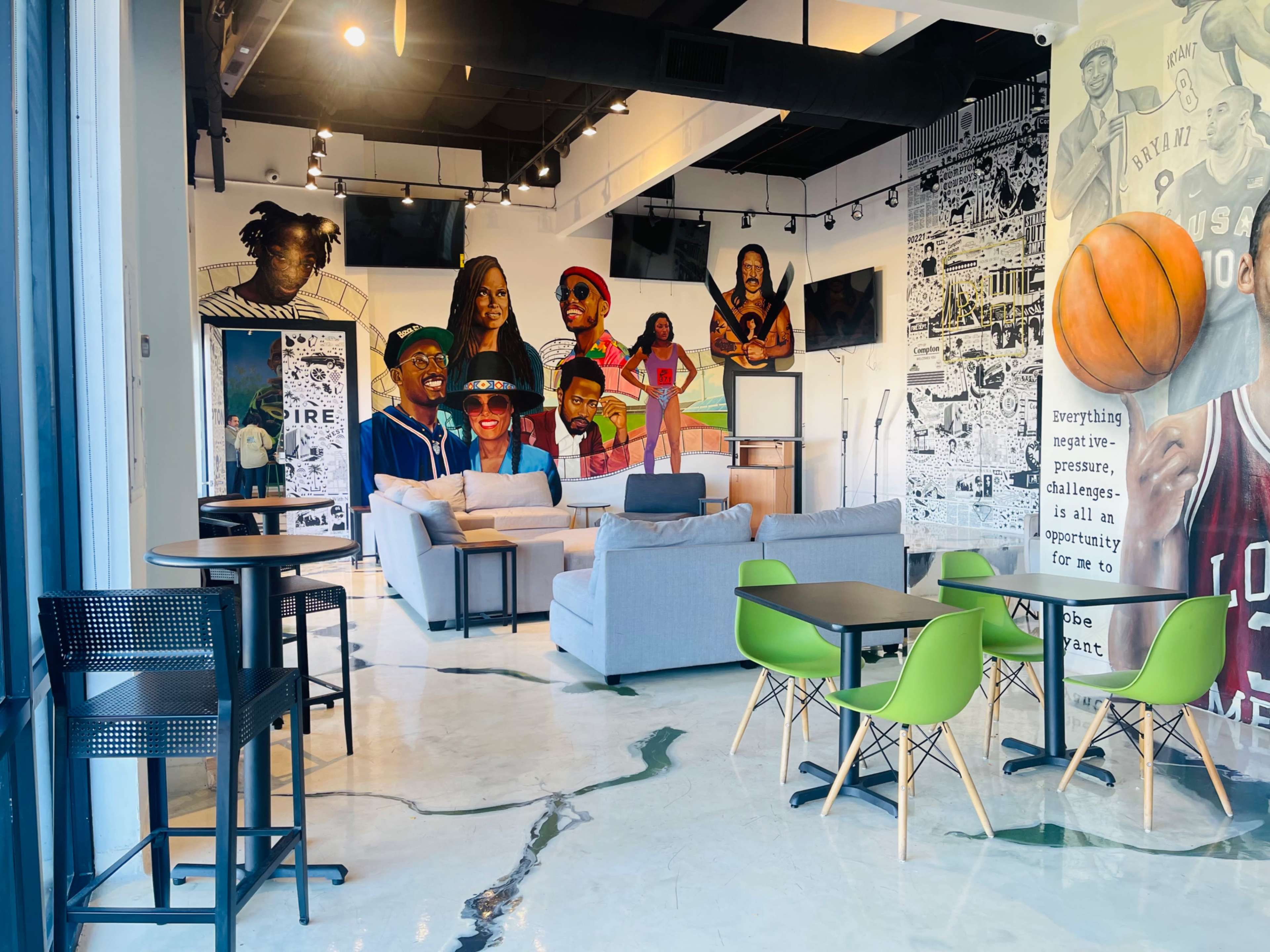 A modern indoor space featuring gray sofas, green chairs, and a large mural with diverse figures, including athletes, on the walls.