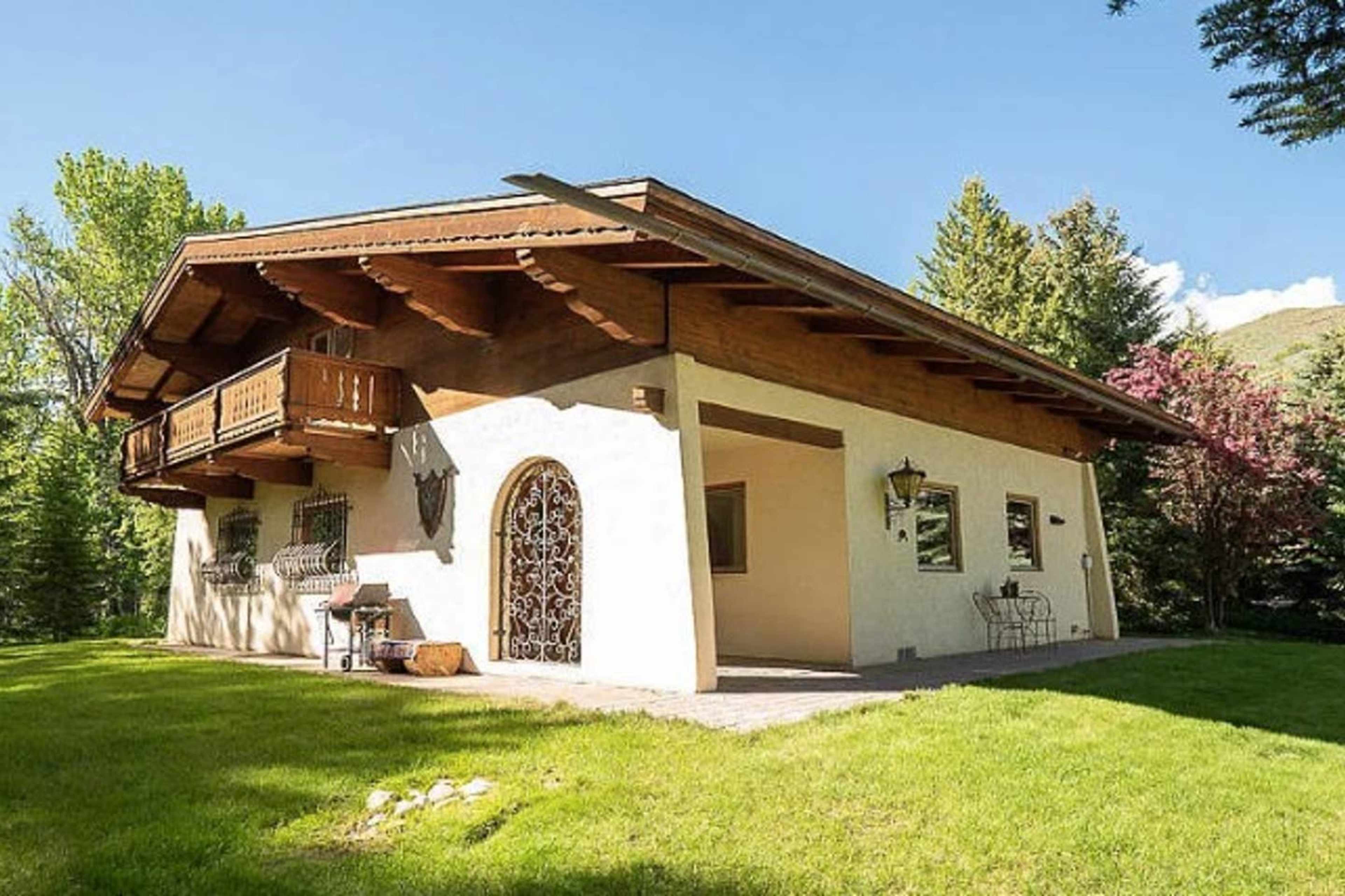 A two-story house with a wooden balcony and decorative iron door is situated on a green lawn surrounded by trees.