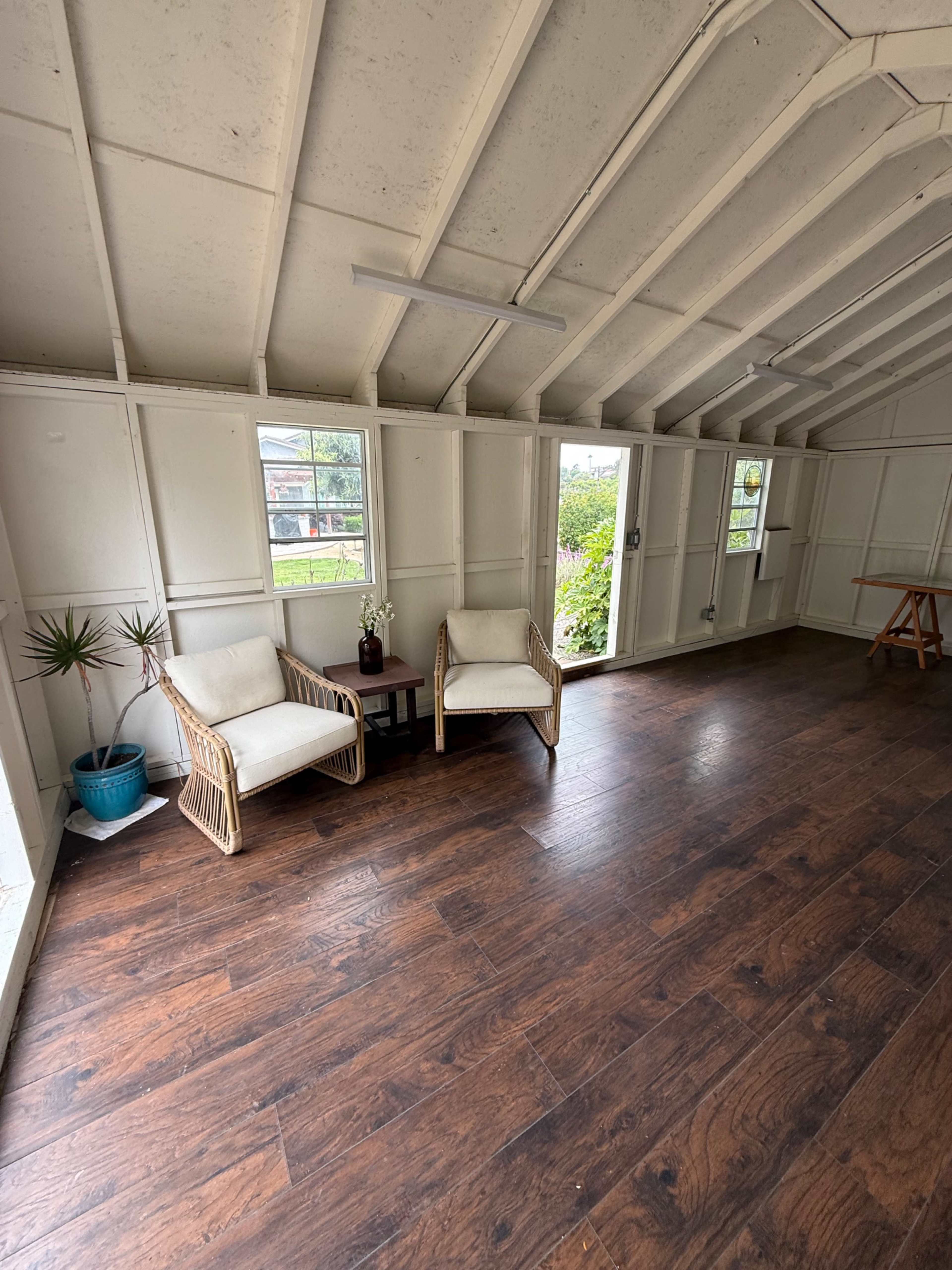 The image shows a spacious room with wood flooring, two light-colored chairs, a small table, and large windows allowing natural light to enter.